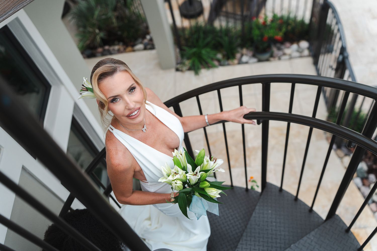 Blonde bride in white gown holds white lily bouquet while descending a spiral staircase outdoors.