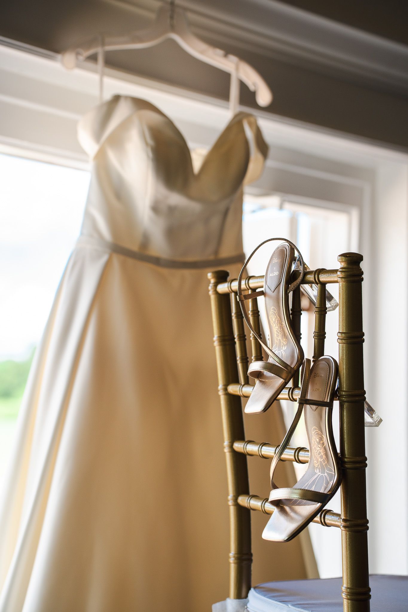 Champagne wedding dress hanging near a window with gold strappy bridal heels displayed on a chair.