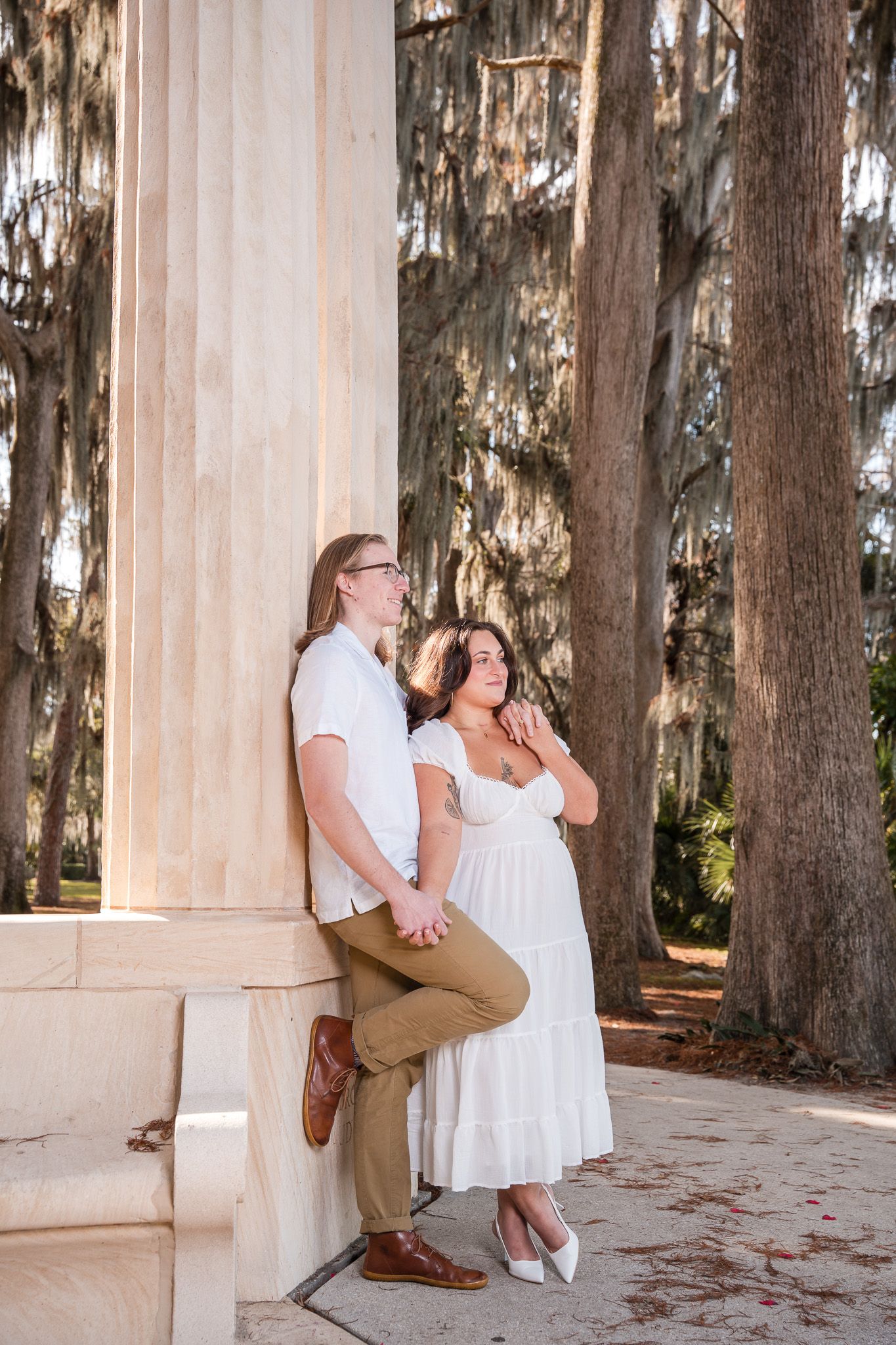 Couple leans against white stone column beneath Spanish moss-draped trees during golden hour engagement session.