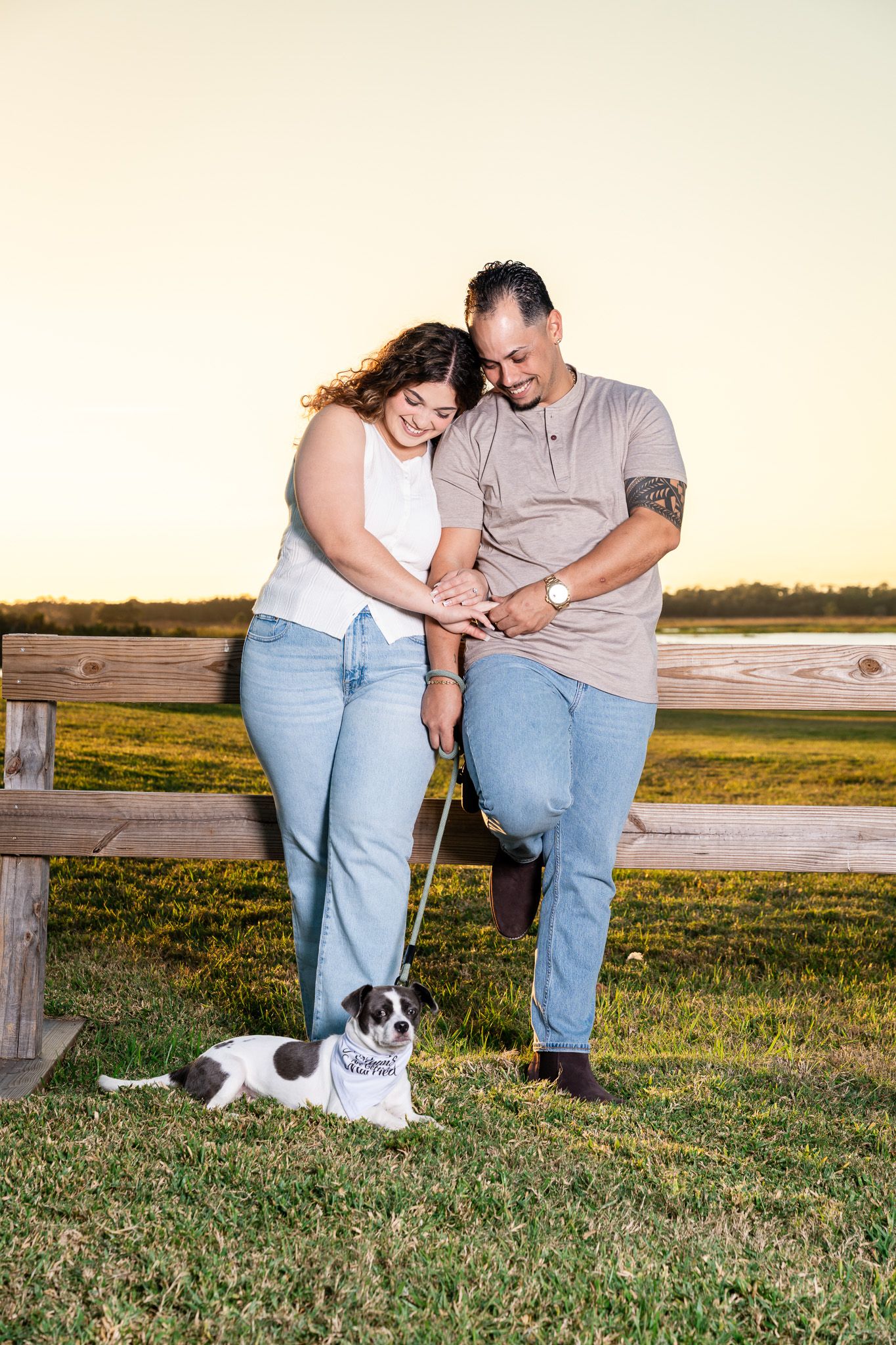 Engaged couple smiling down at their small dog on a leash near a wooden fence at sunset.