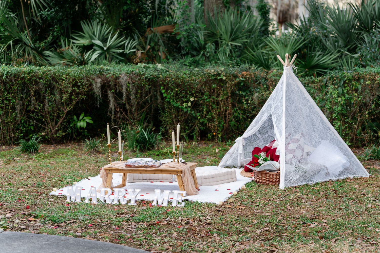 Romantic outdoor proposal setup with white teepee tent, picnic table, candles and rose petals.