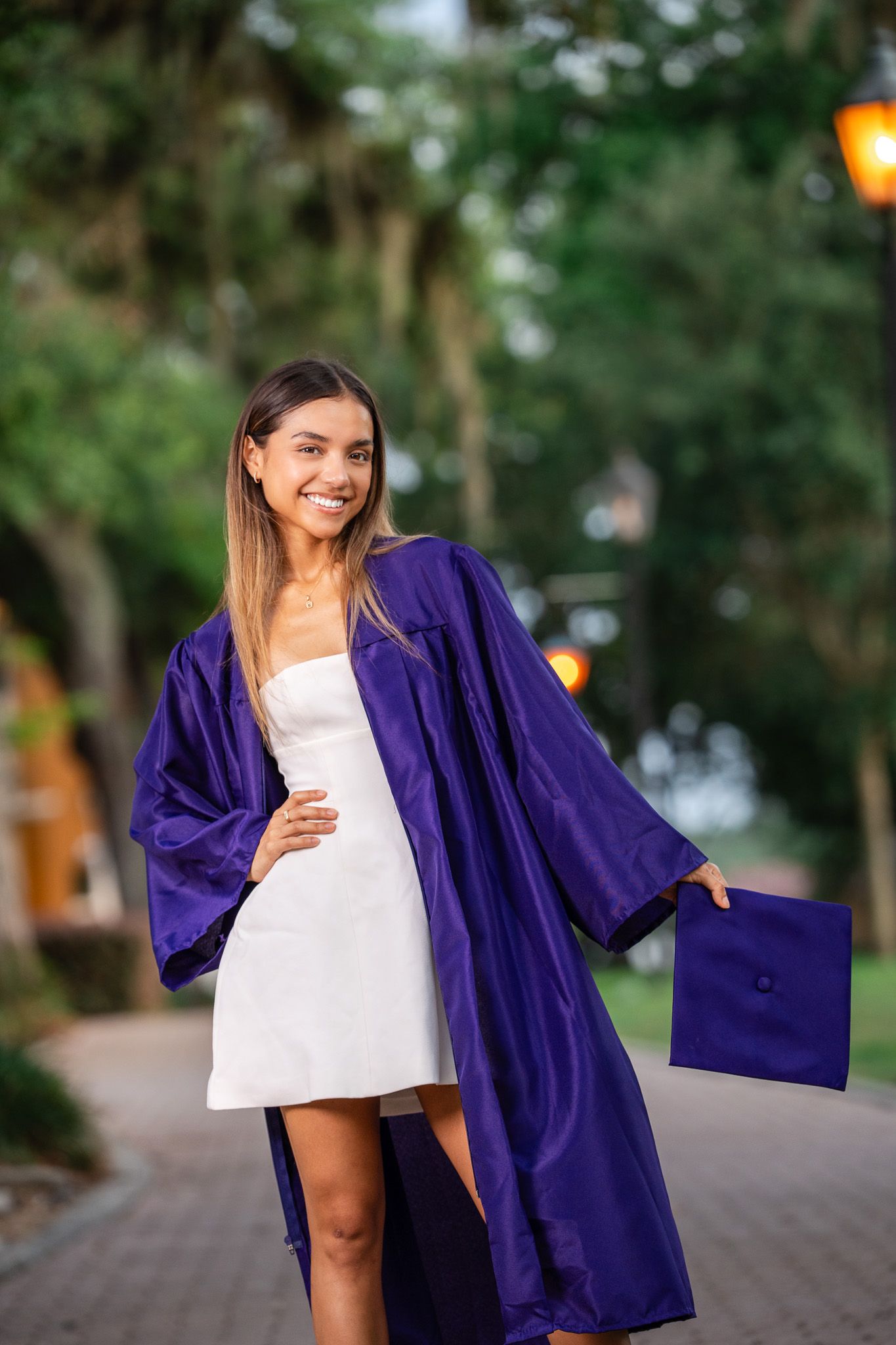 Happy female graduate in purple cap and gown smiles holding her cap on a scenic brick walkway.