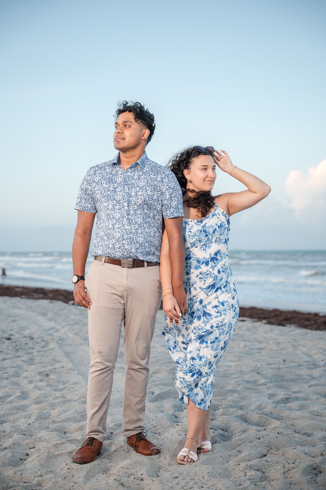 Couple in matching blue floral outfits holding hands on a sandy beach at dusk with ocean waves in the background.