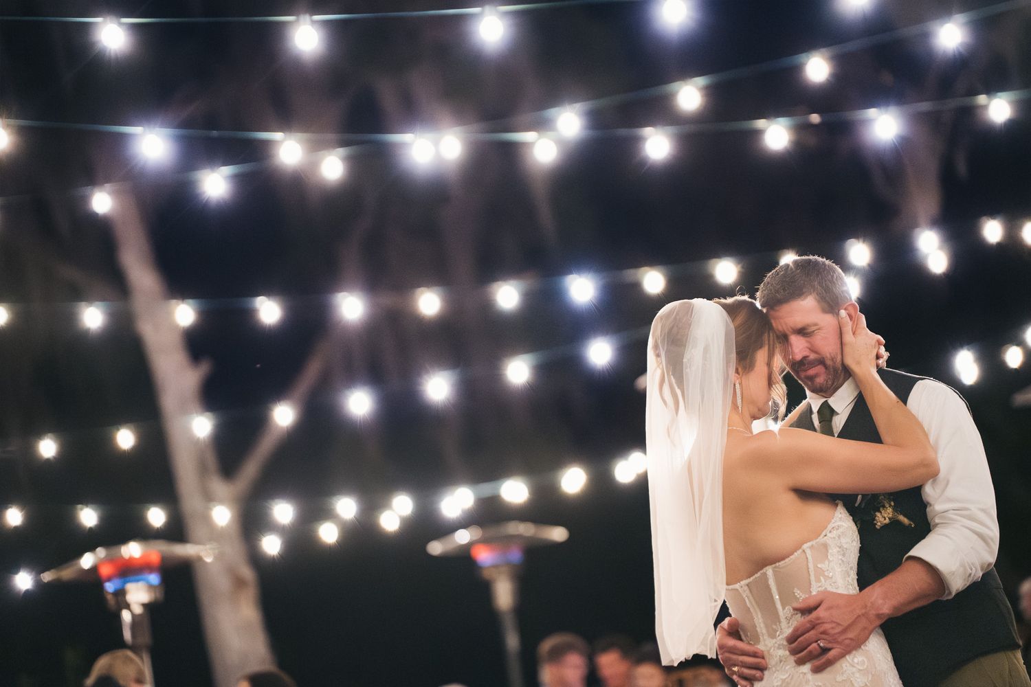 Bride and groom sharing first dance under romantic string lights at outdoor evening wedding reception.