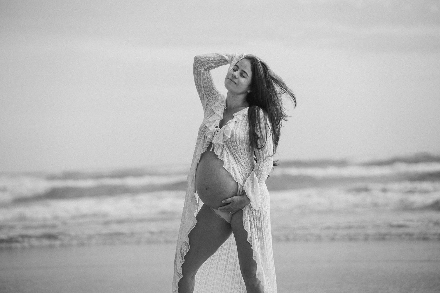 Pregnant woman in white dress posing gracefully on beach in black and white photo.