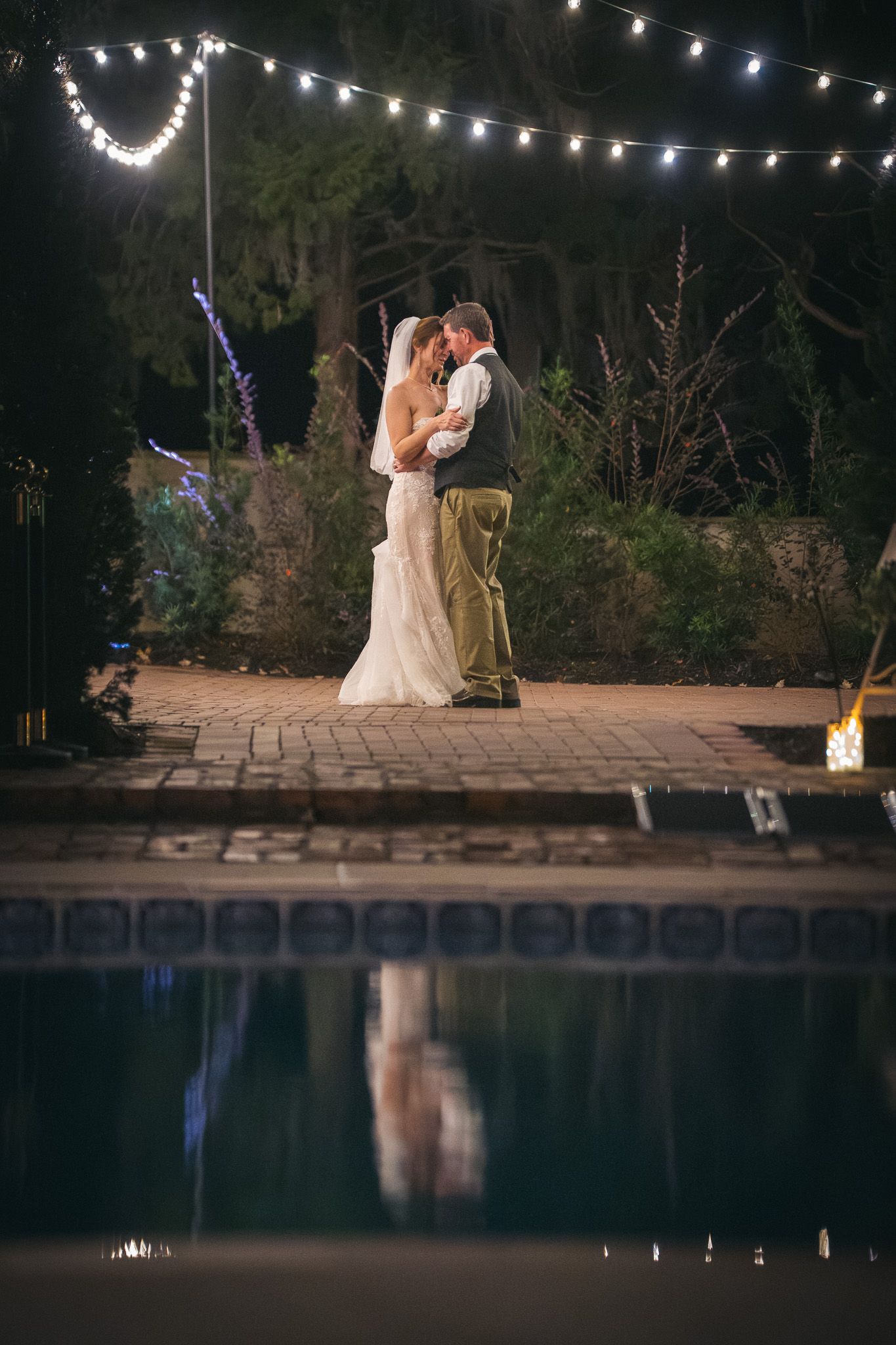 Bride and groom share a romantic embrace by a pool at night under twinkling string lights.