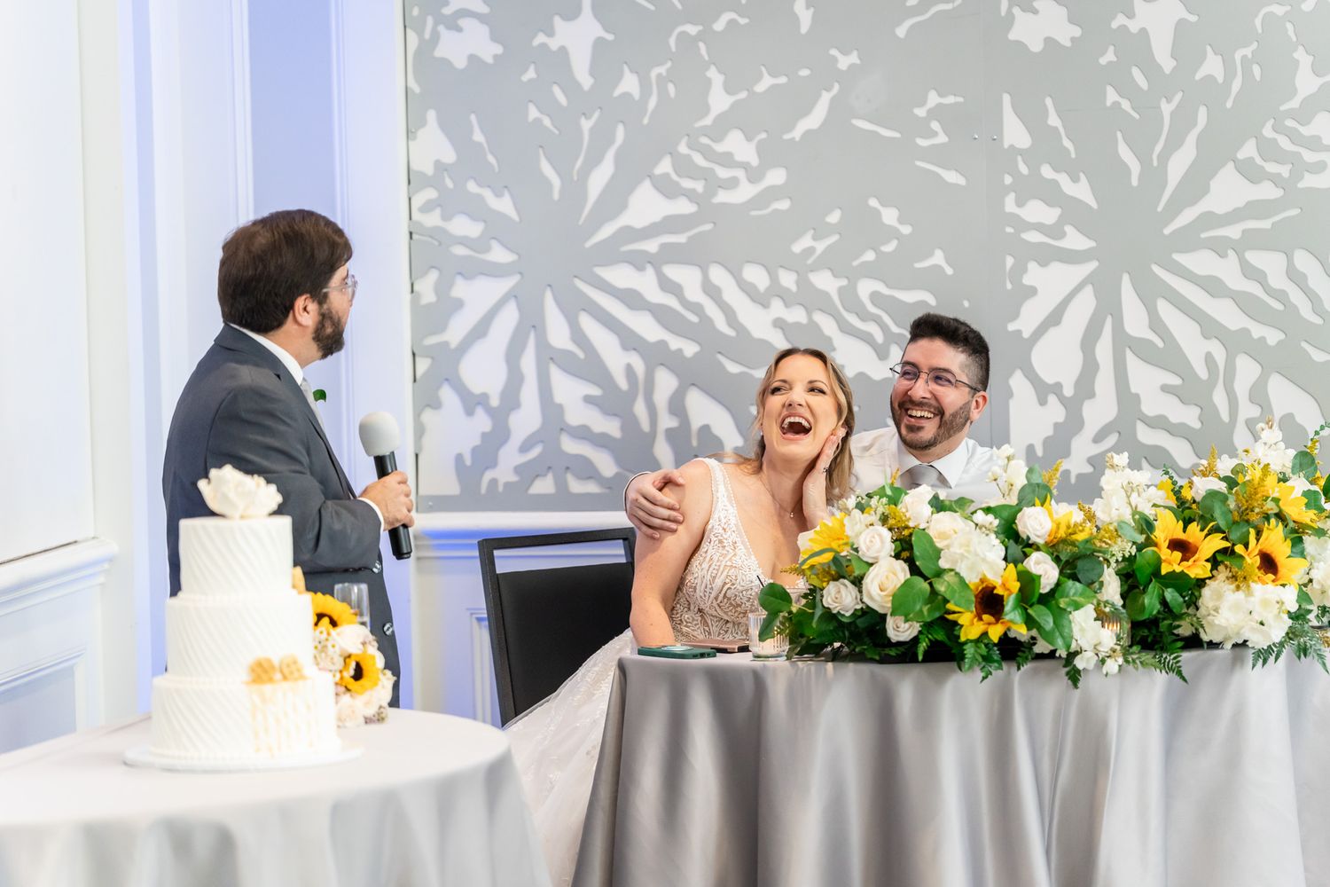 Bride and groom laugh joyfully during wedding toast at decorated sweetheart table.