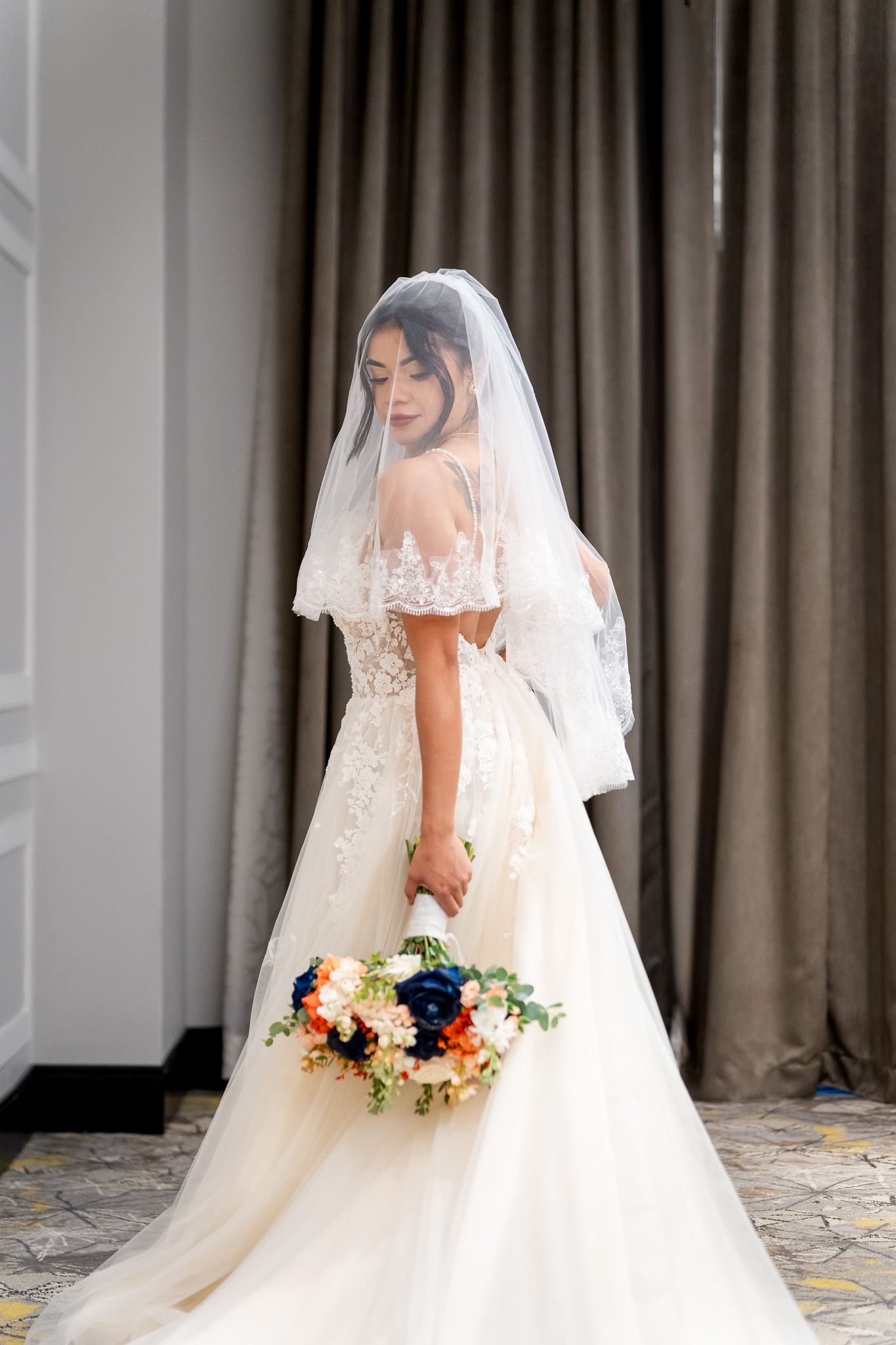 Elegant bride in lace wedding gown with veil holds colorful bouquet of blue and orange flowers.