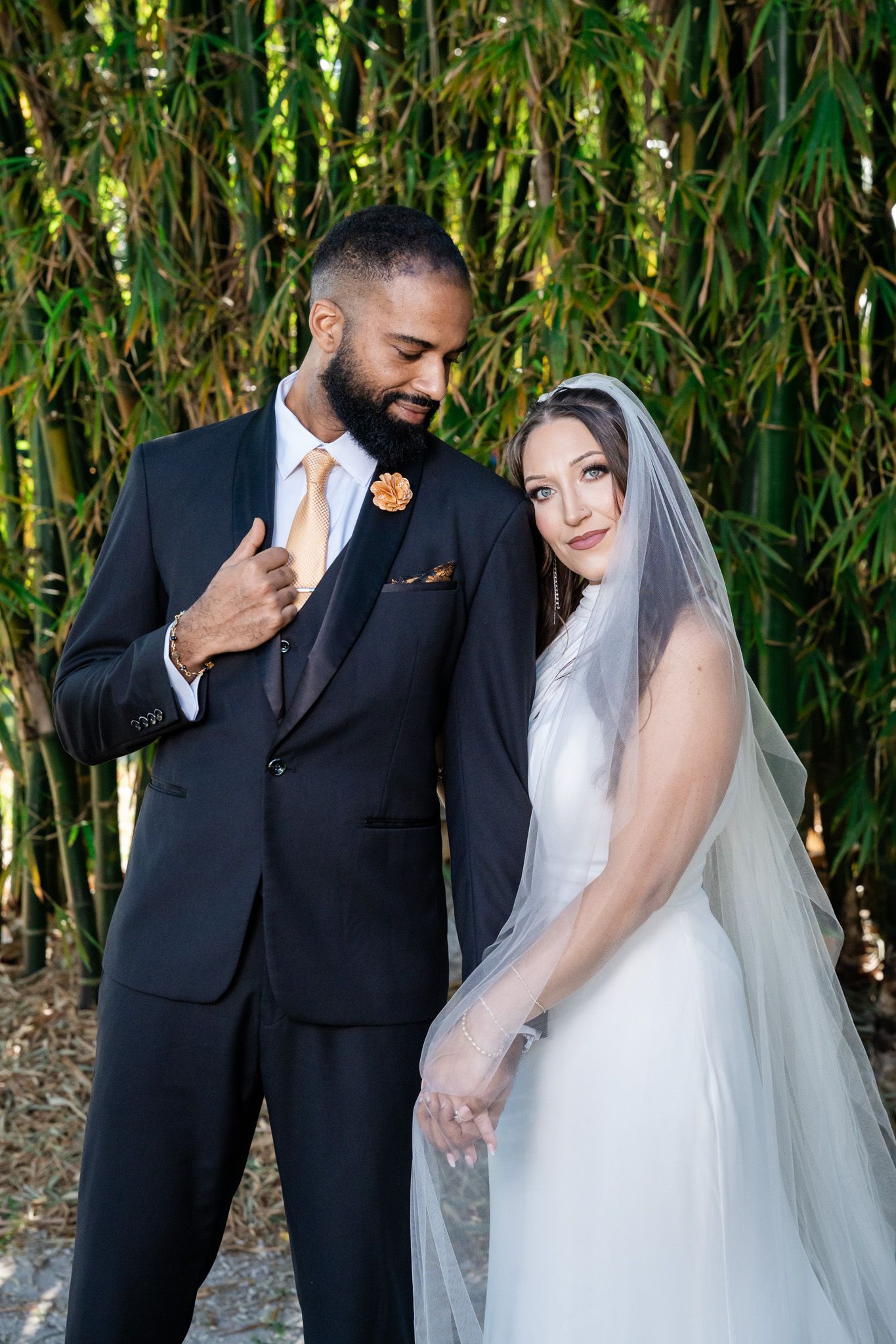 Bride in white veil leans on groom in black suit as they pose tenderly before bamboo greenery.