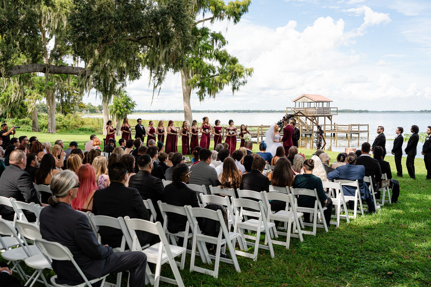 Outdoor lakeside wedding ceremony with bridal party in burgundy dresses and seated guests.