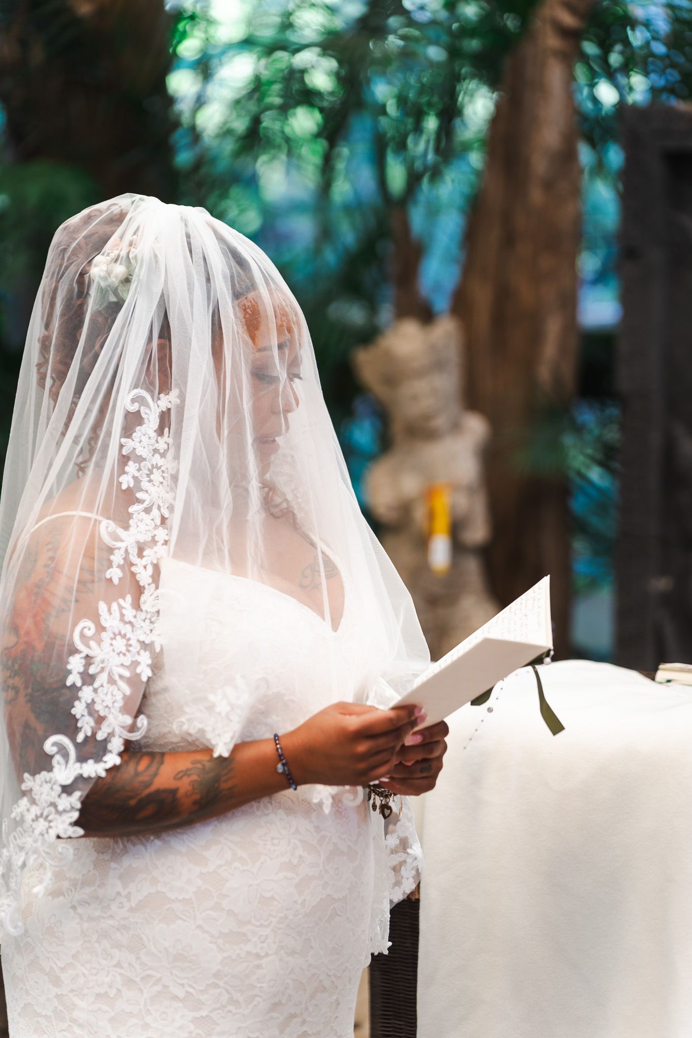 Bride in lace wedding gown and veil reads vows from paper during outdoor ceremony.