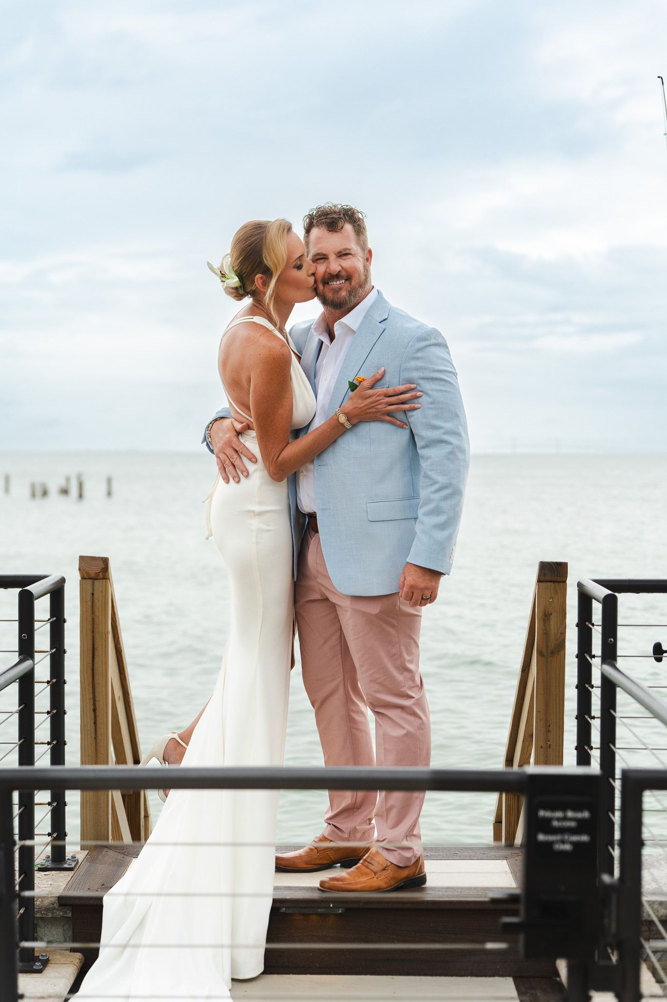 Bride kisses groom on cheek while posing on waterfront dock with ocean backdrop on wedding day.