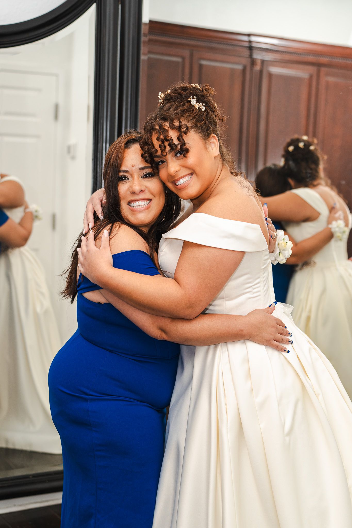 Bride in off-shoulder white gown shares warm embrace with bridesmaid in royal blue dress.