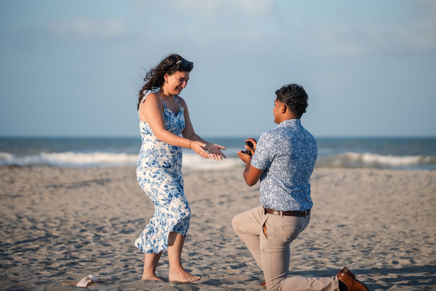 Man proposing on one knee to surprised woman on a sandy beach, both dressed in blue floral attire.