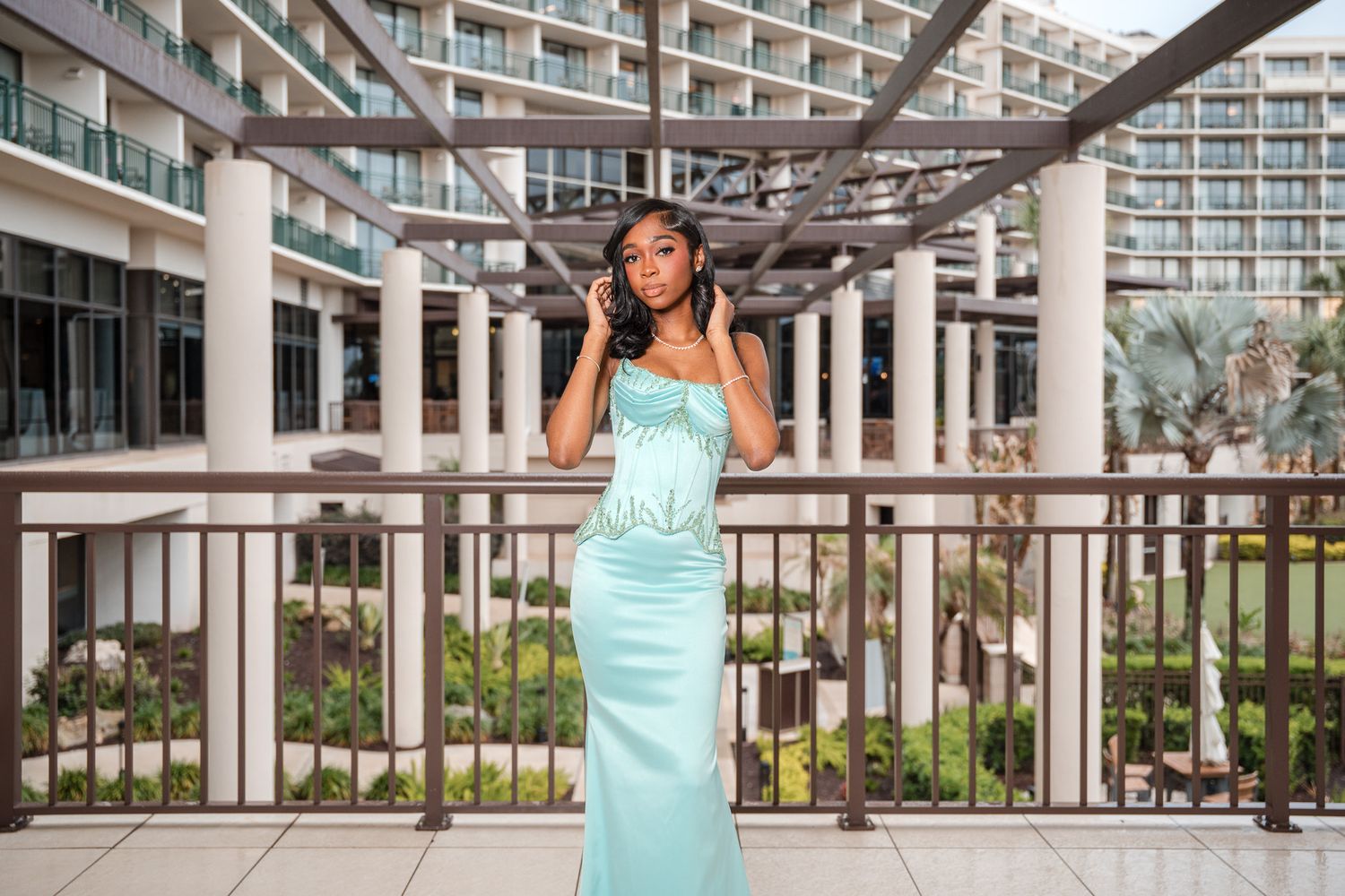 A woman poses in a elegant mint green strapless gown on a balcony with a luxury hotel backdrop.