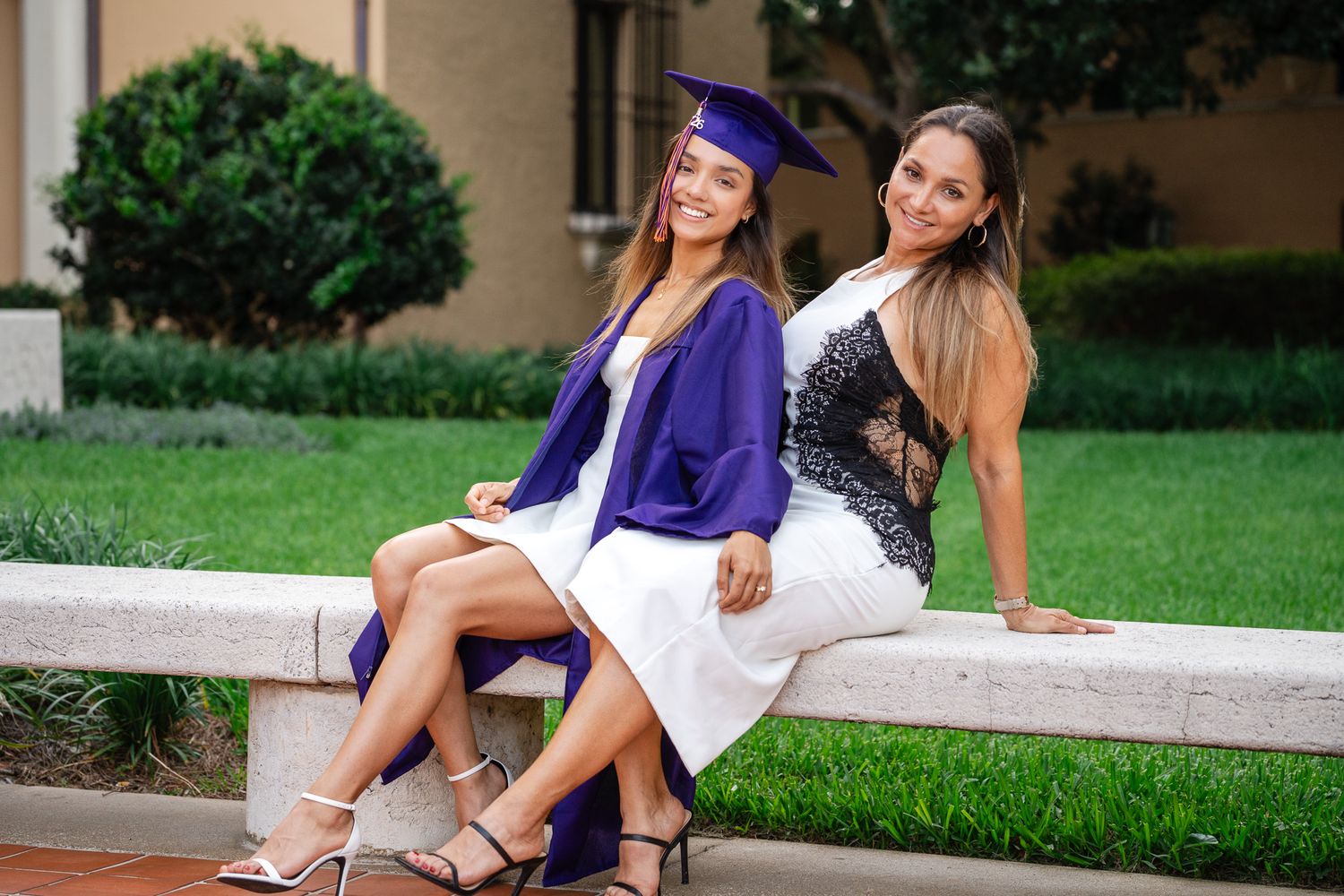 Two young women smile sitting outdoors, one wearing purple graduation cap and gown, celebrating academic achievement.