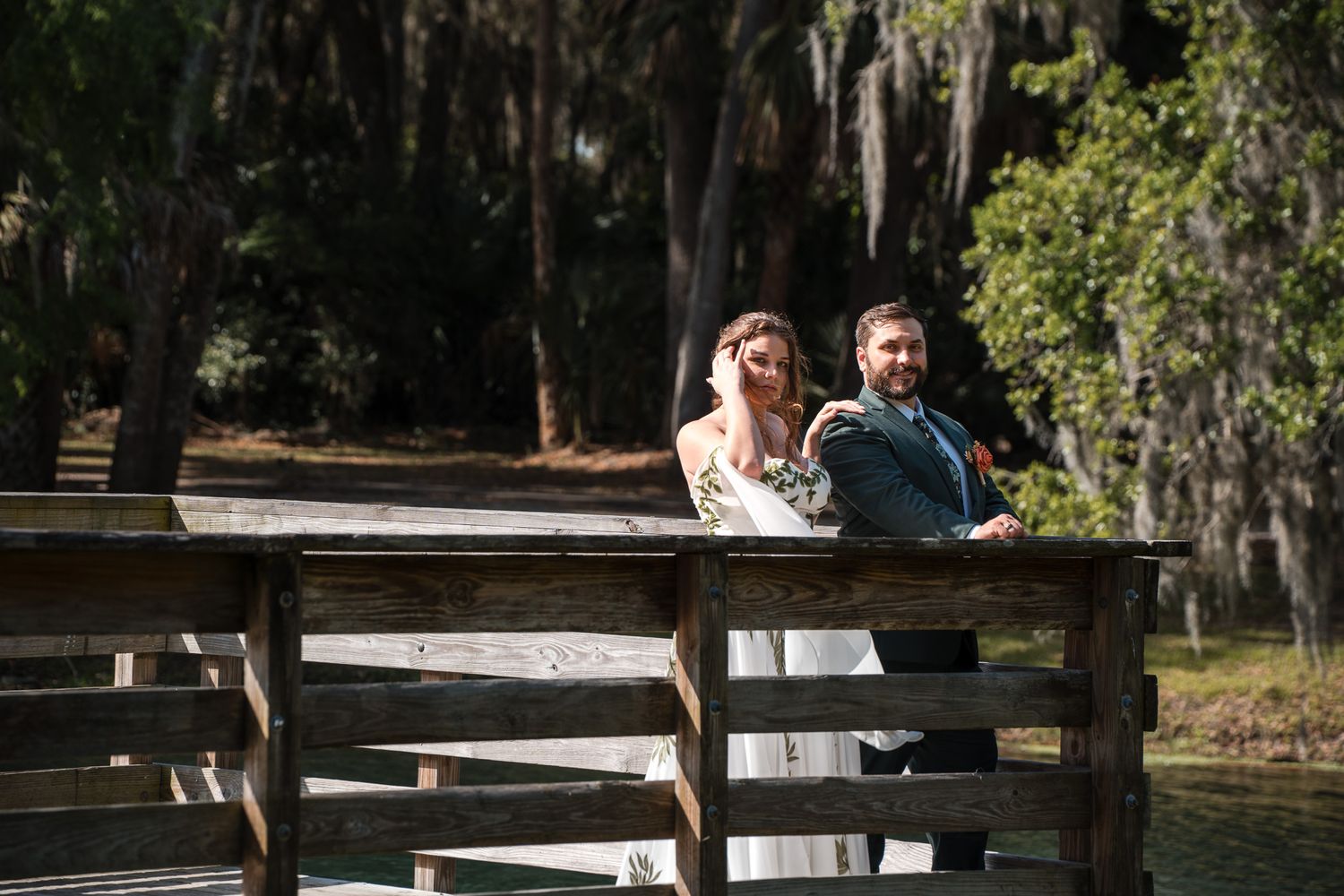 A bride and groom lean against a wooden dock railing surrounded by lush trees in a romantic outdoor wedding photo.