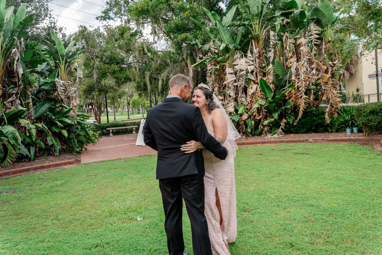Bride and groom share a romantic first look moment on lush green lawn surrounded by tropical trees at outdoor wedding venue.
