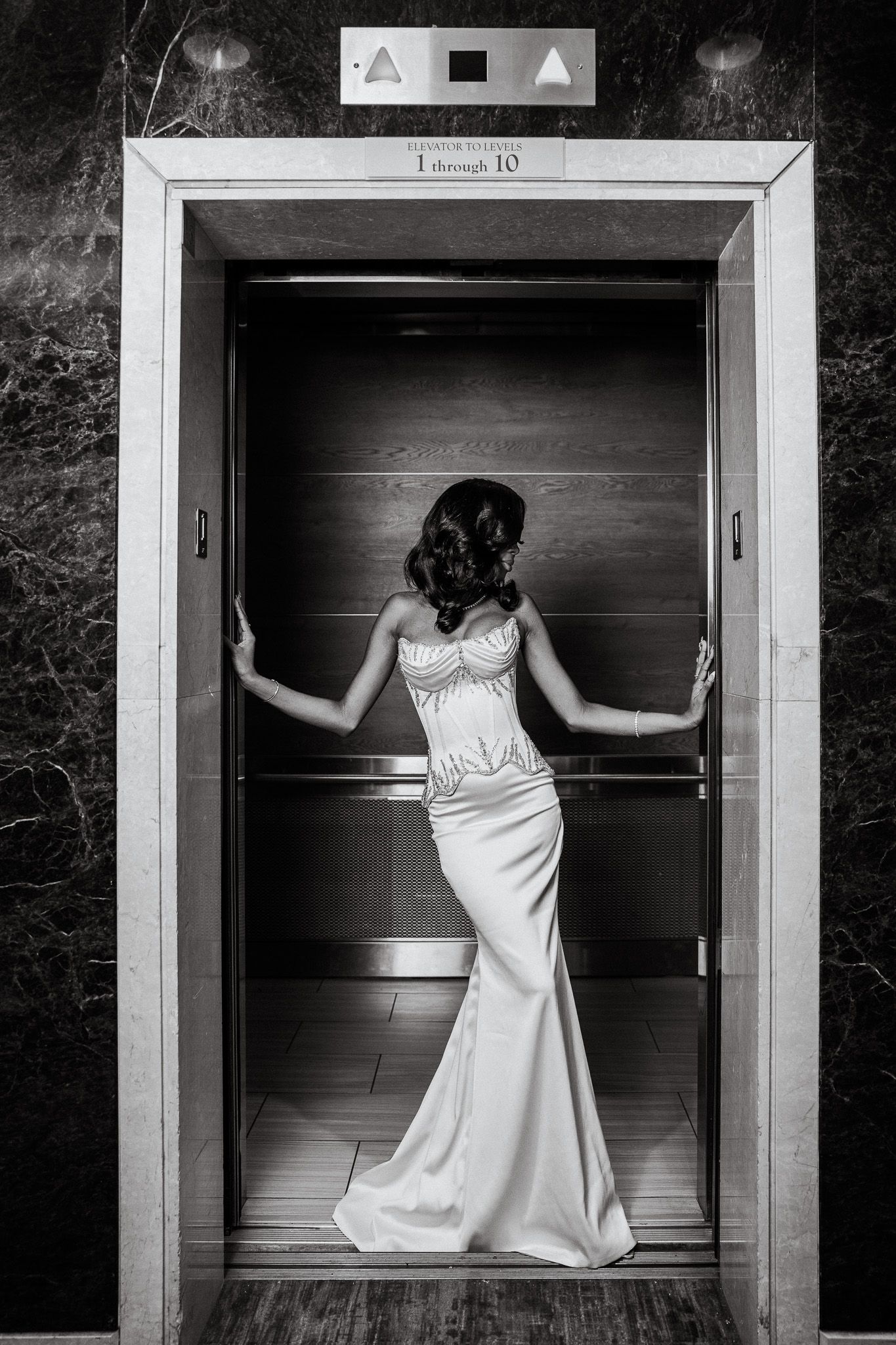 Elegant woman in white strapless gown posing inside elevator doors, black and white fashion photography.
