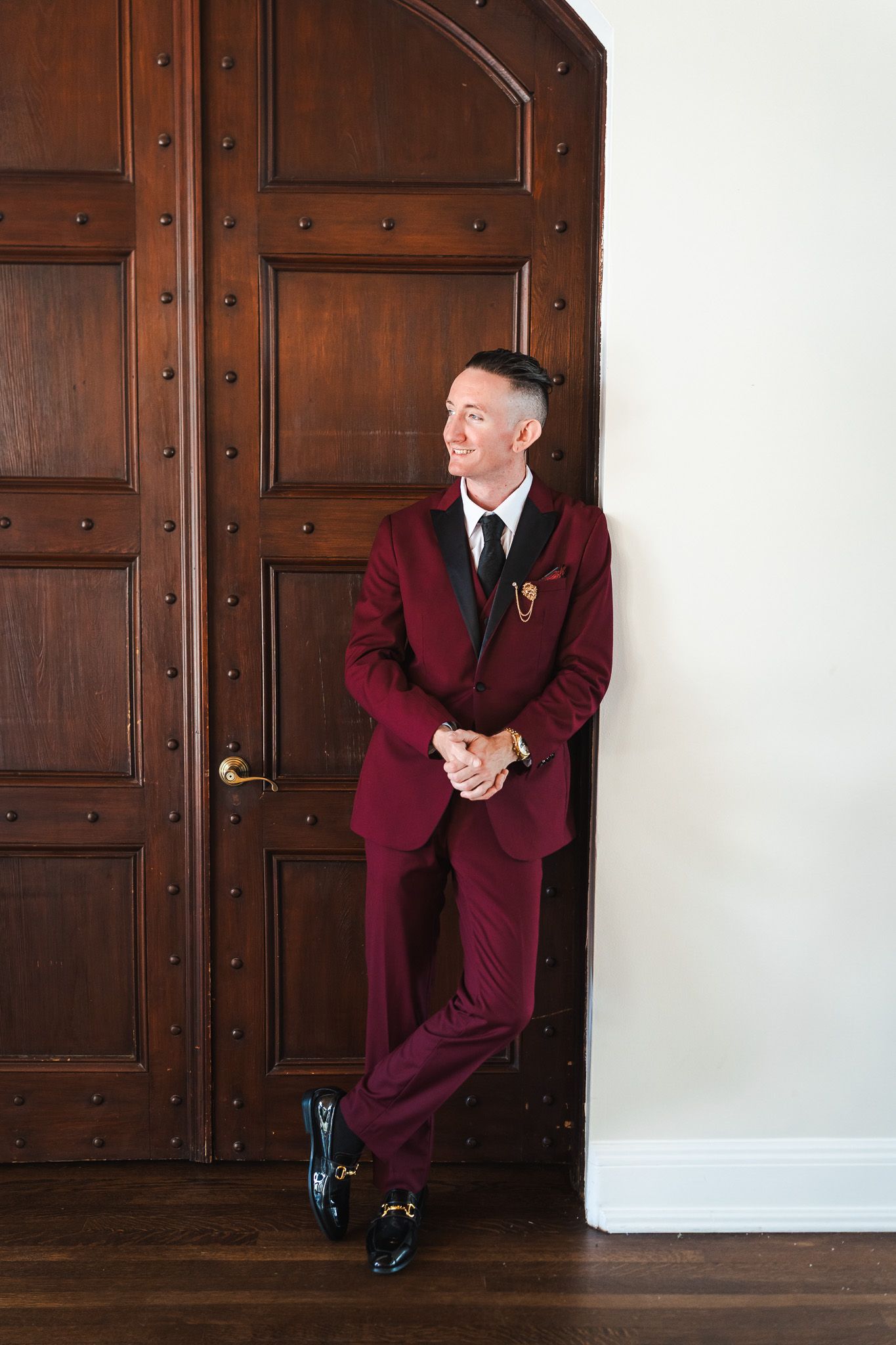 Dapper groom in burgundy suit and black tie leans against ornate wooden doors smiling.
