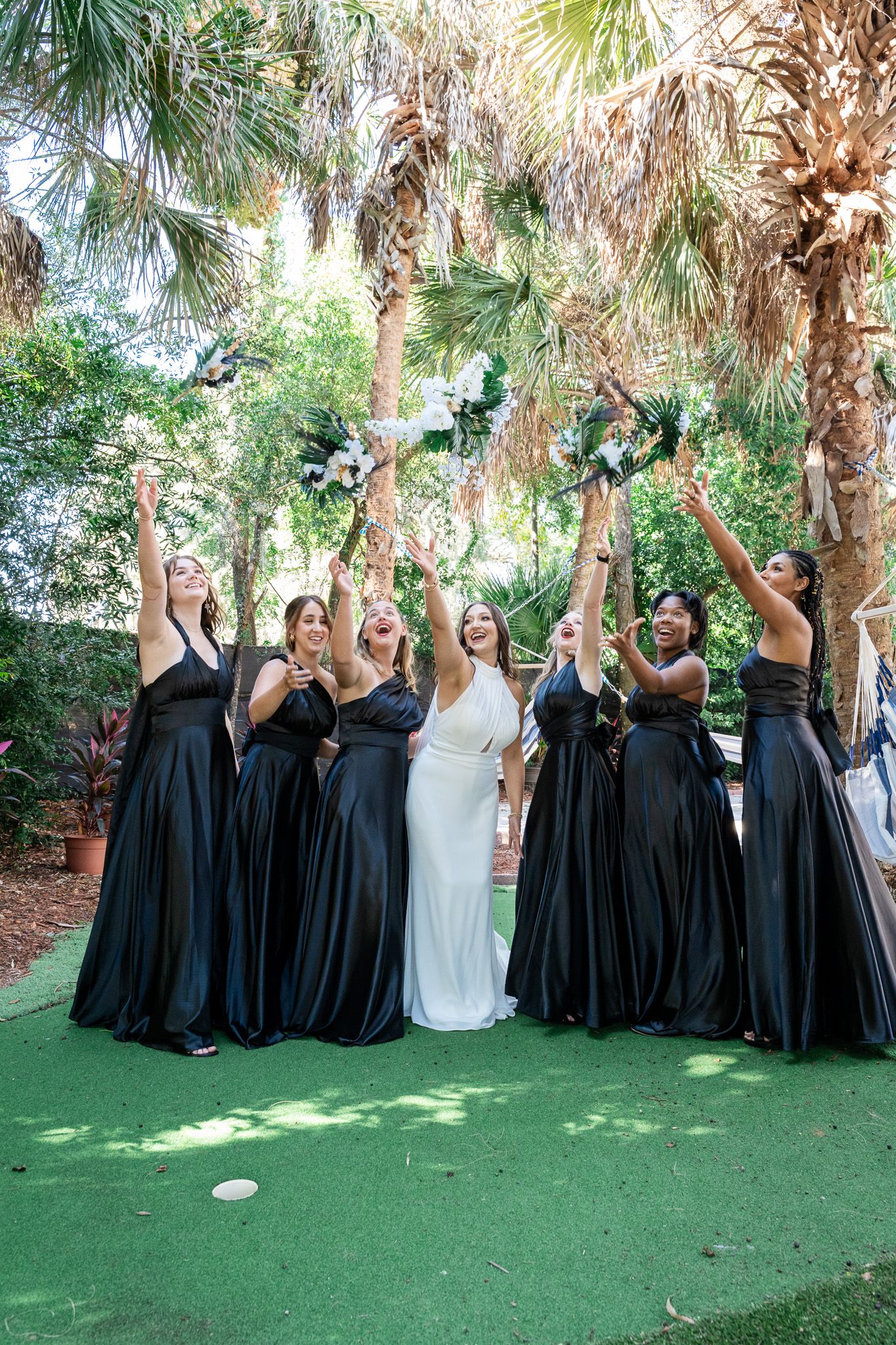 Bridesmaids in black gowns raise bouquets joyfully with bride in white dress near palm trees.