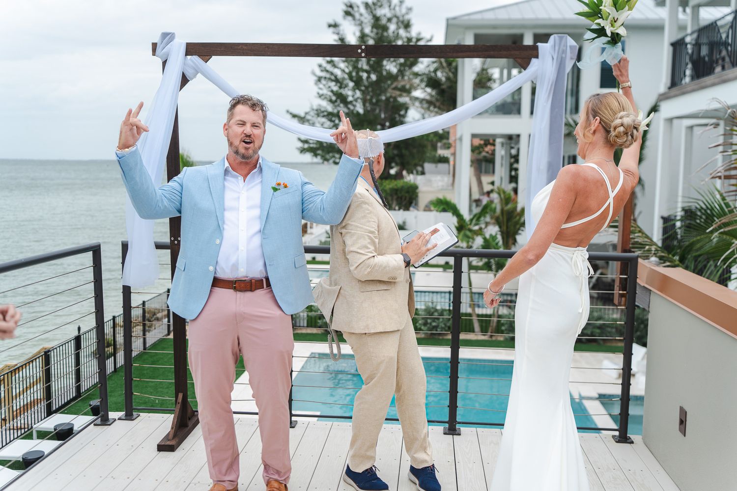 Excited groom in light blue blazer cheers as bride approaches outdoor beachside wedding altar.