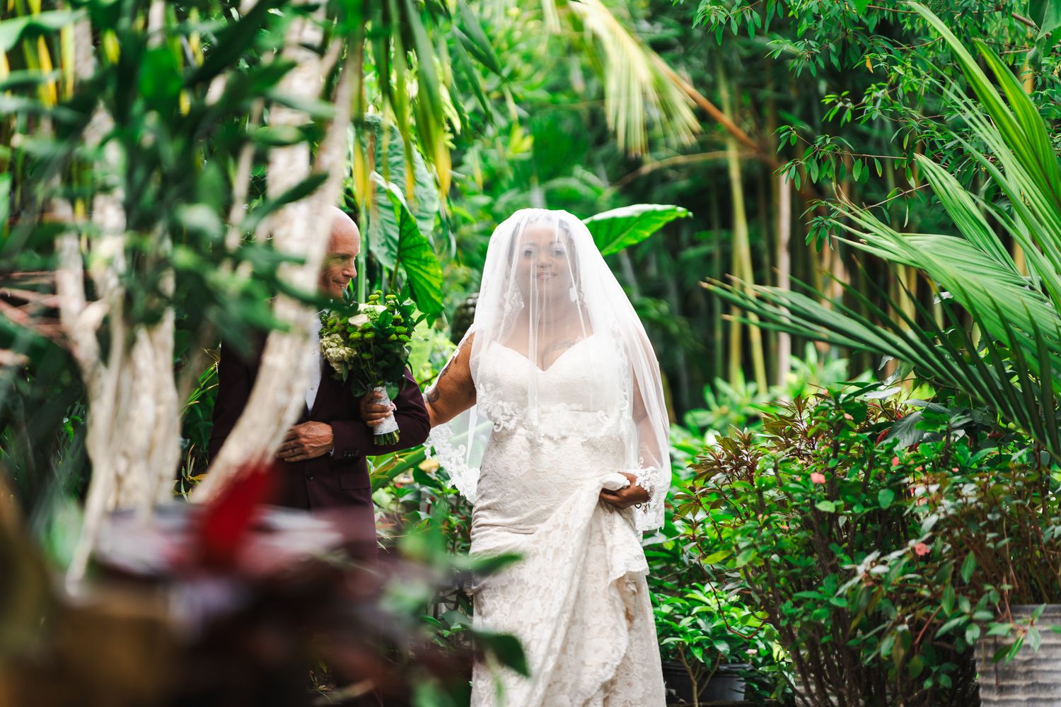 Bride in white lace gown and veil being escorted through a vibrant tropical garden ceremony path.