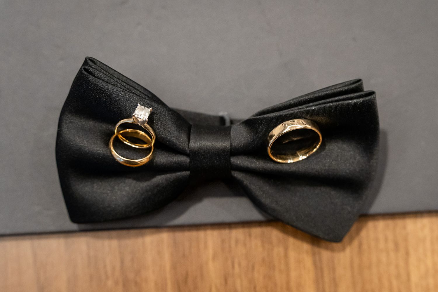 Two gold wedding bands and a diamond engagement ring displayed on a black satin bow tie.