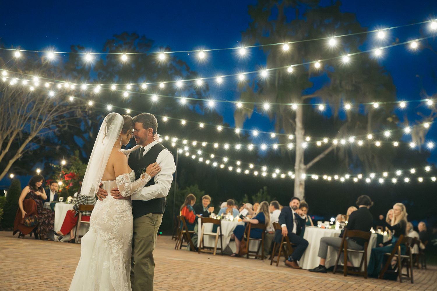 Bride and groom share their first dance outdoors under glowing string lights with guests seated nearby.