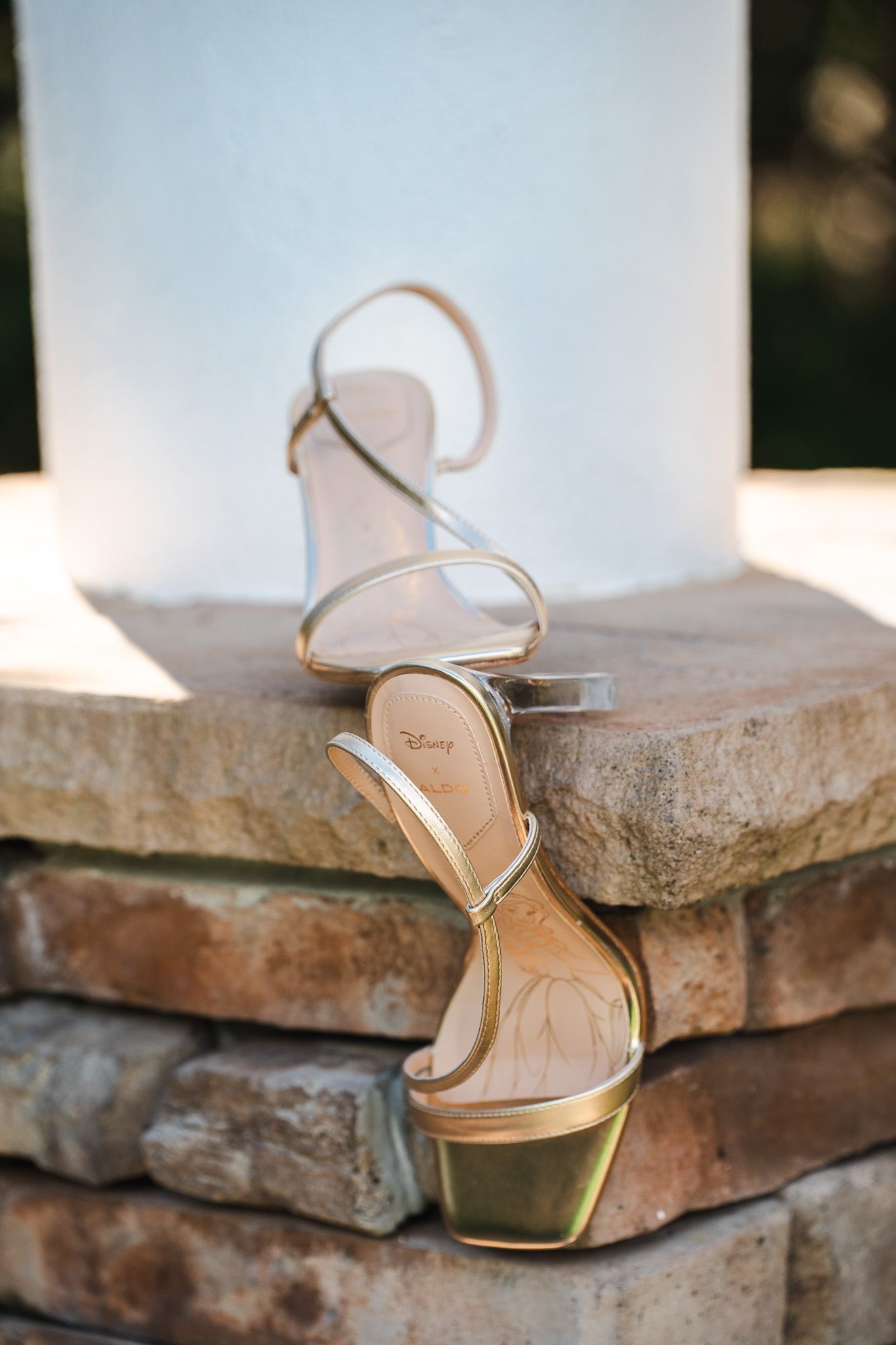 Gold Disney strappy bridal heels displayed on stone pillar in outdoor wedding setting.