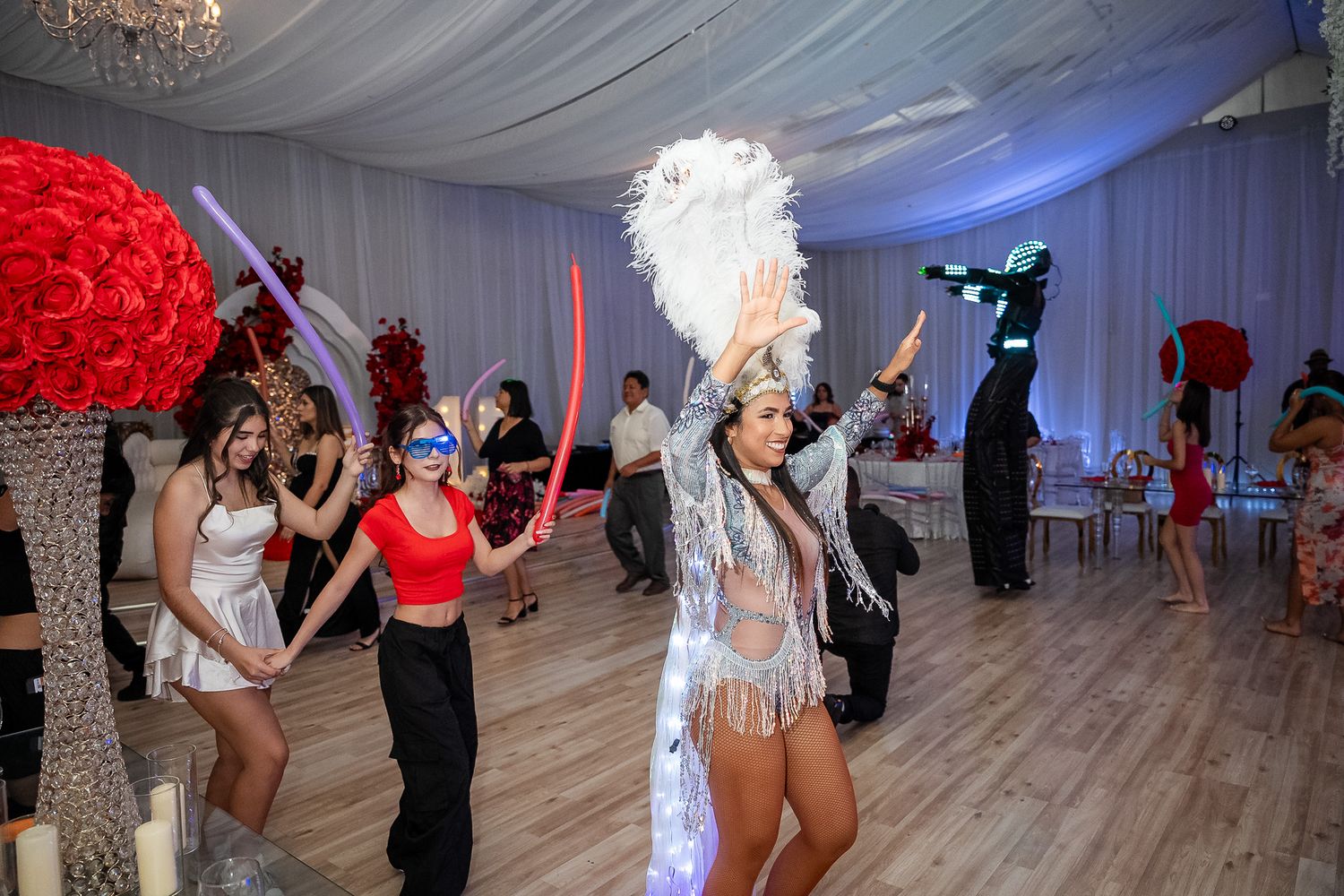 Energetic quinceañera party performers dance with LED props and feathered headdress in decorated white draped ballroom.