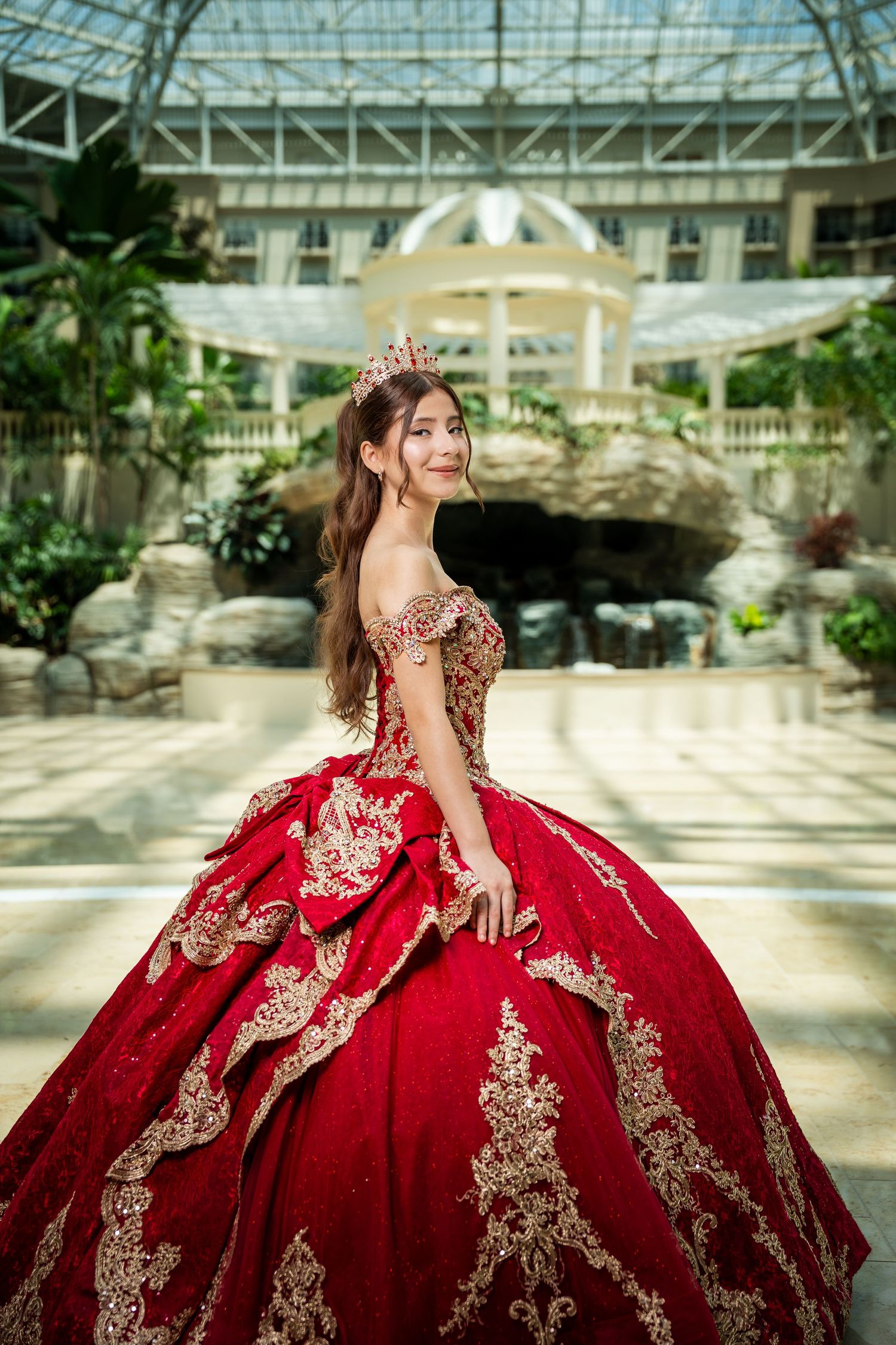Young woman in red and gold quinceañera ball gown with crown poses in elegant glass atrium.