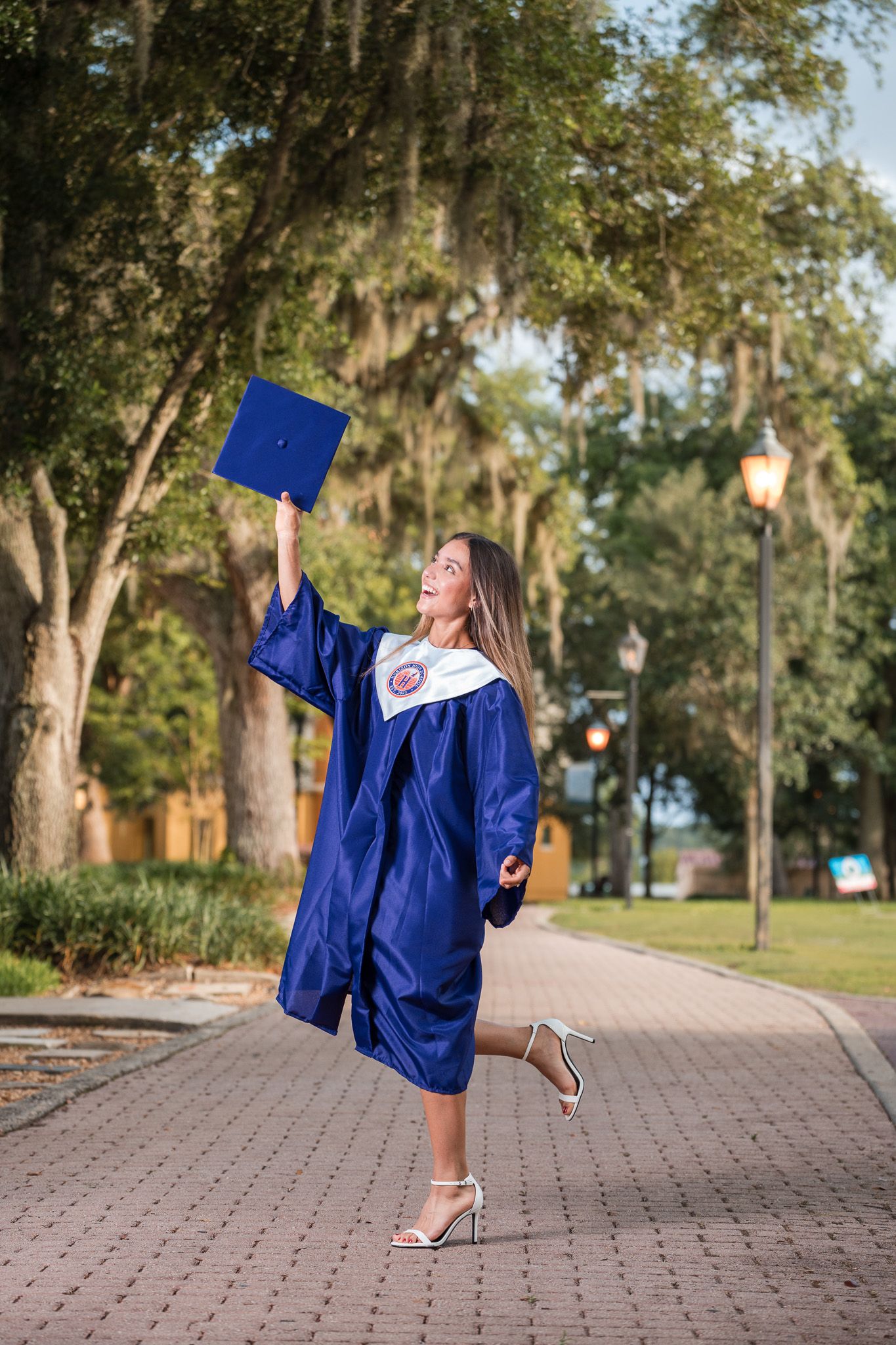 Young woman in blue graduation gown raises cap triumphantly on a tree-lined brick pathway.