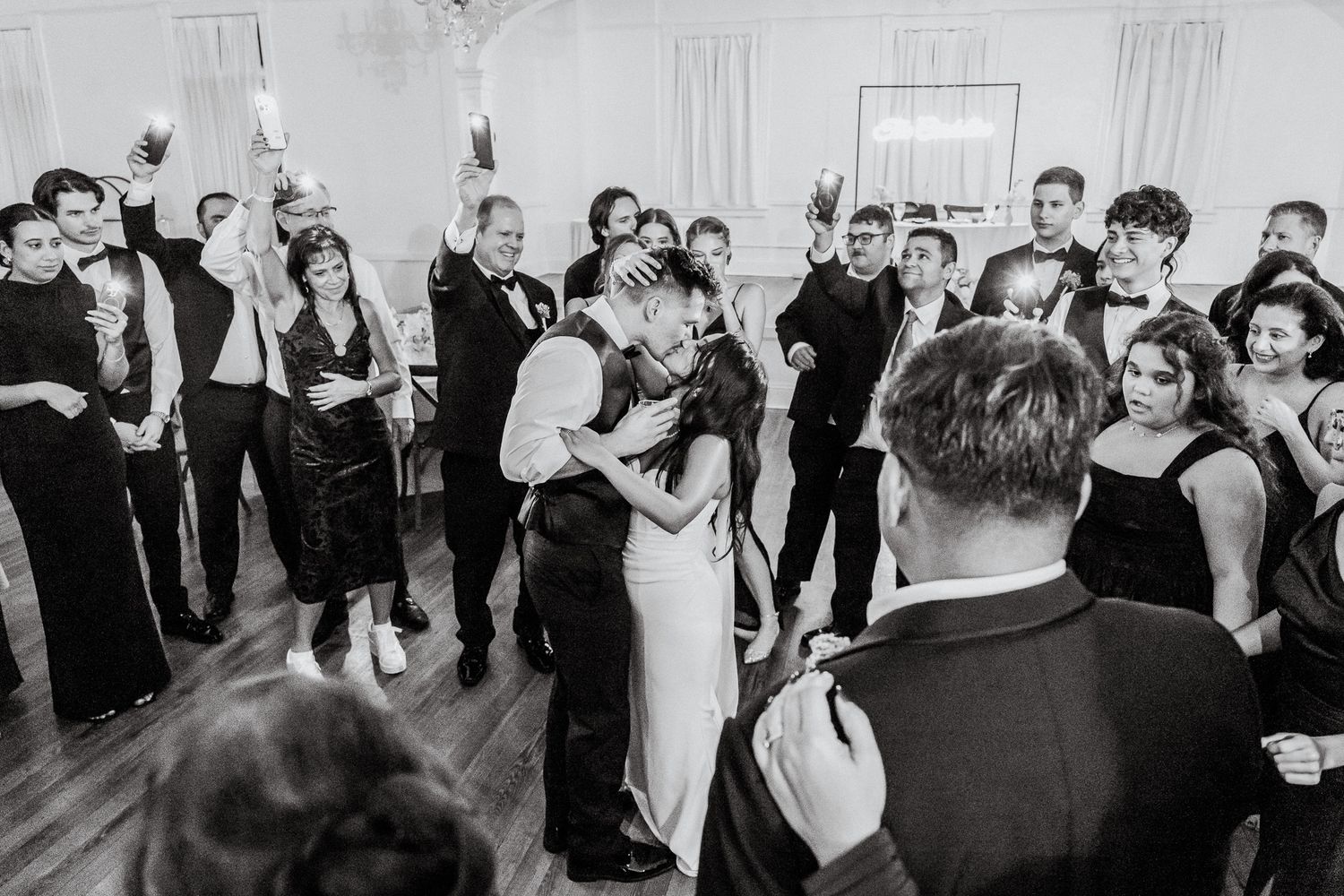 Bride and groom kissing on dance floor surrounded by cheering wedding guests in black and white photo.