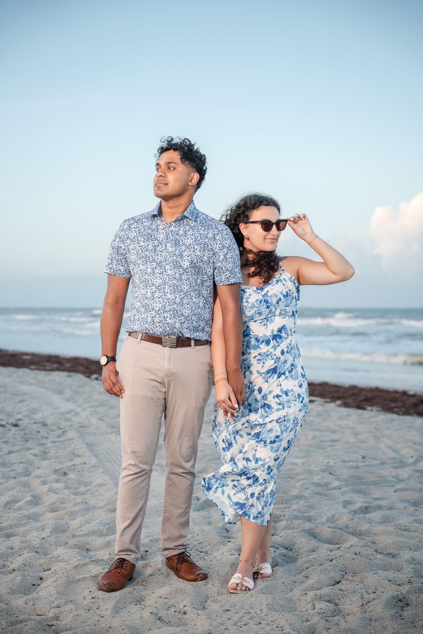 Couple holding hands on a sandy beach at sunset, man in floral shirt and woman in blue floral dress.