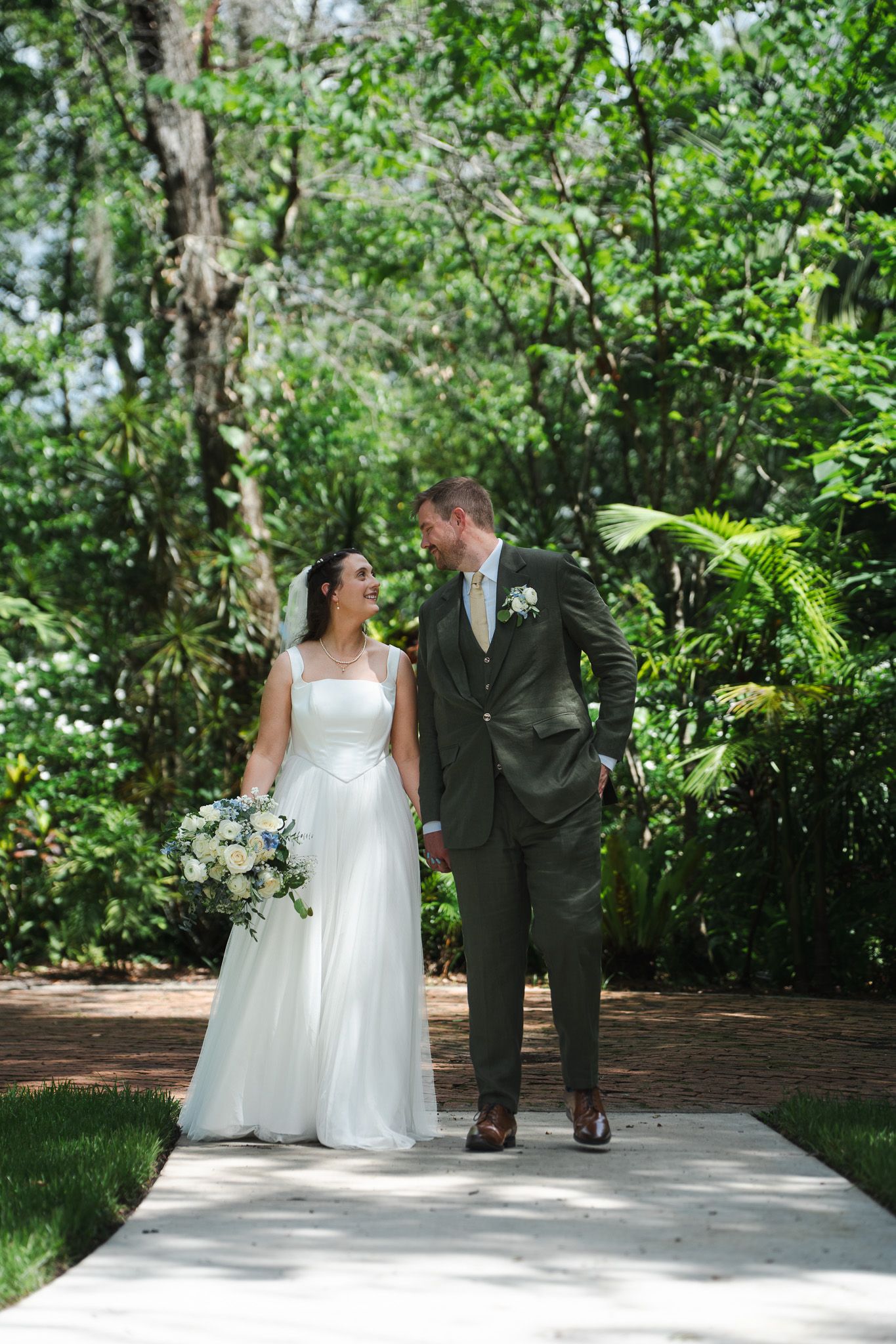 Bride and groom walk hand in hand along a sunlit garden pathway surrounded by lush green trees.