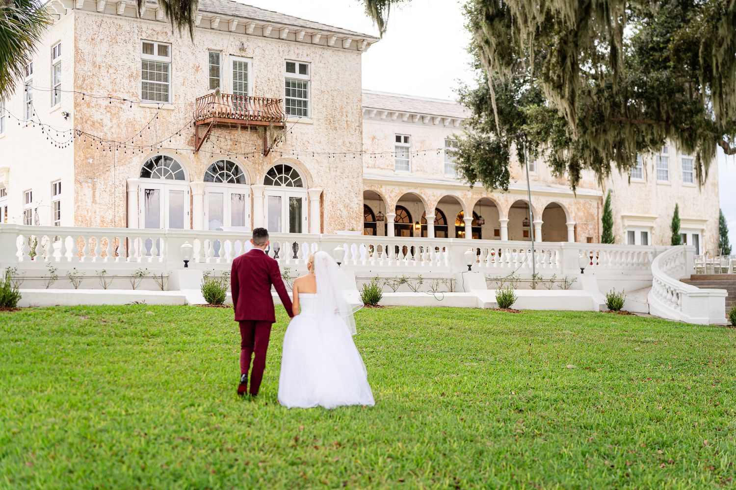 Bride and groom walking hand in hand toward a grand historic mansion surrounded by lush greenery.