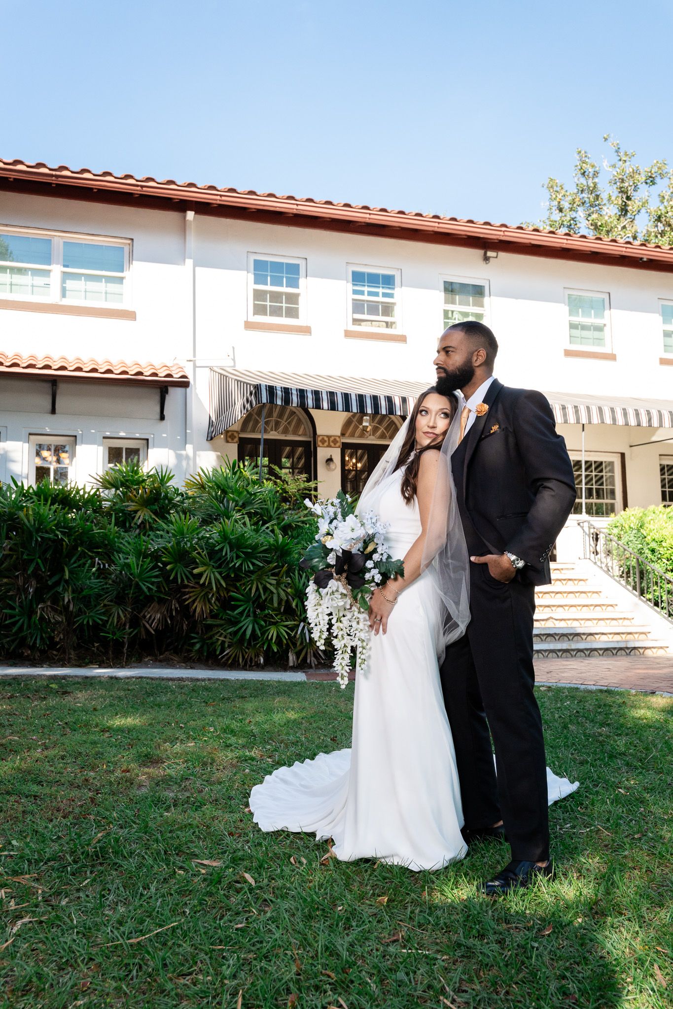 Bride and groom pose together on lawn in front of elegant white Spanish-style wedding venue building.