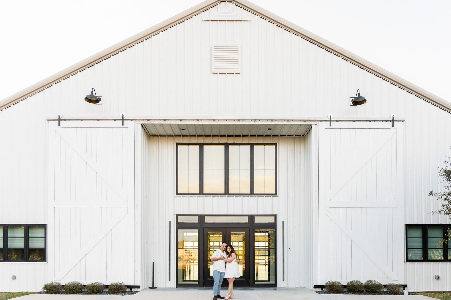 Couple embracing in front of a large white barn venue with black-framed doors at golden hour.