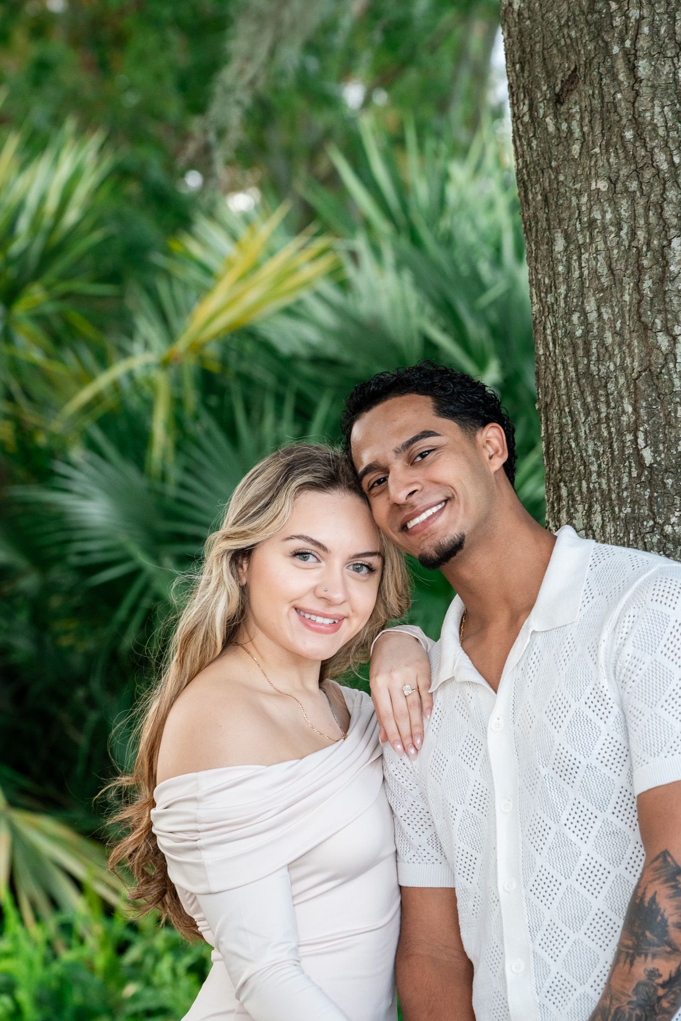 Smiling interracial couple leaning against tree surrounded by lush tropical palm foliage.