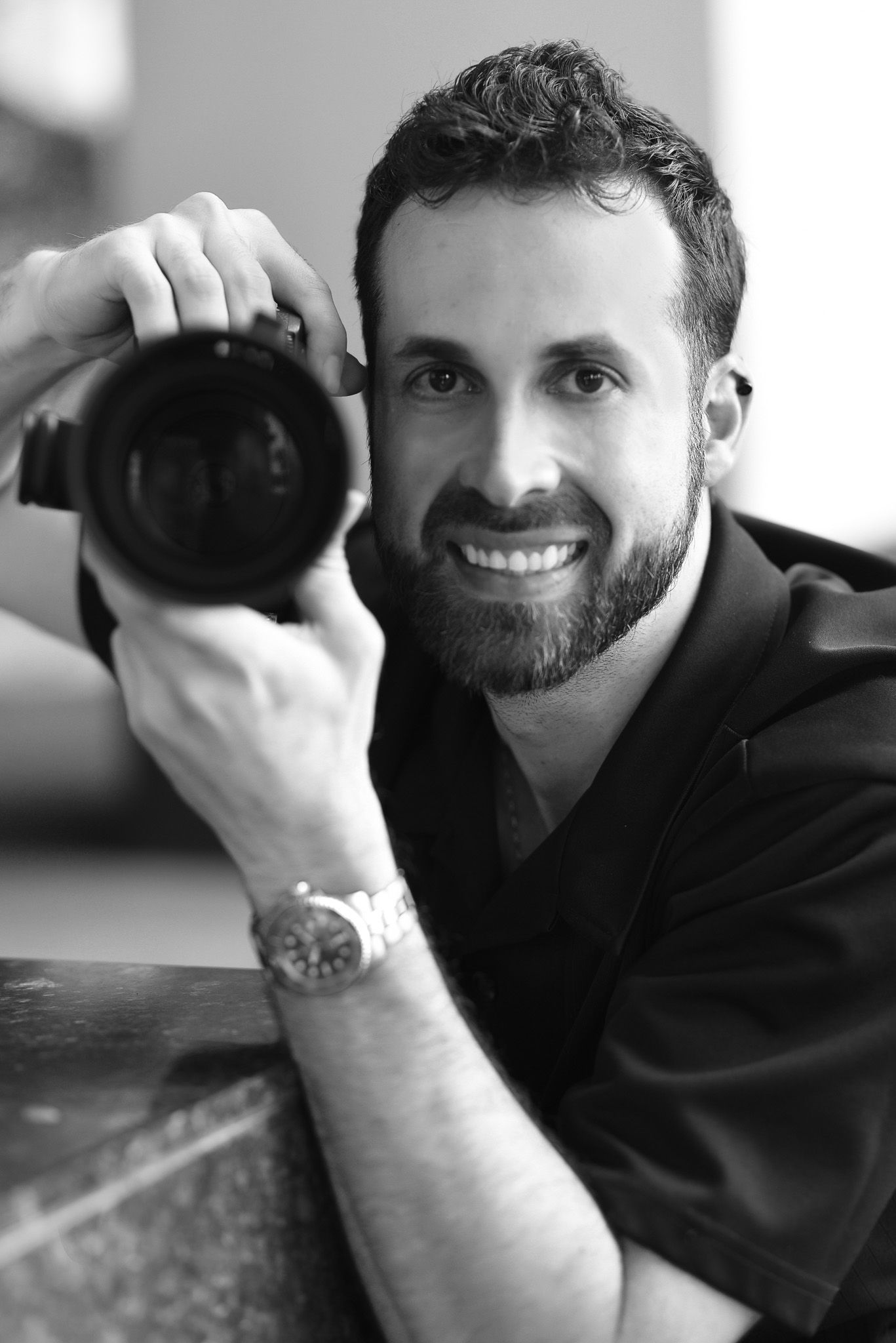 Smiling bearded man holding a DSLR camera in a black and white portrait photo.