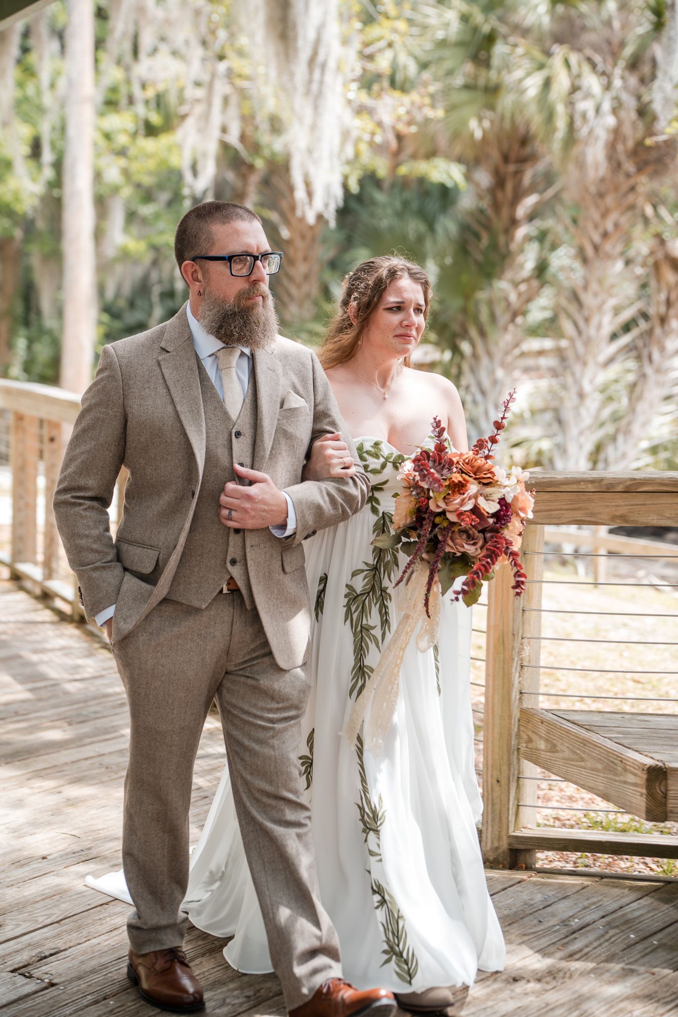 Groom in tan tweed suit and bride in white gown with autumn bouquet walk arm-in-arm on a sunlit wooden bridge.