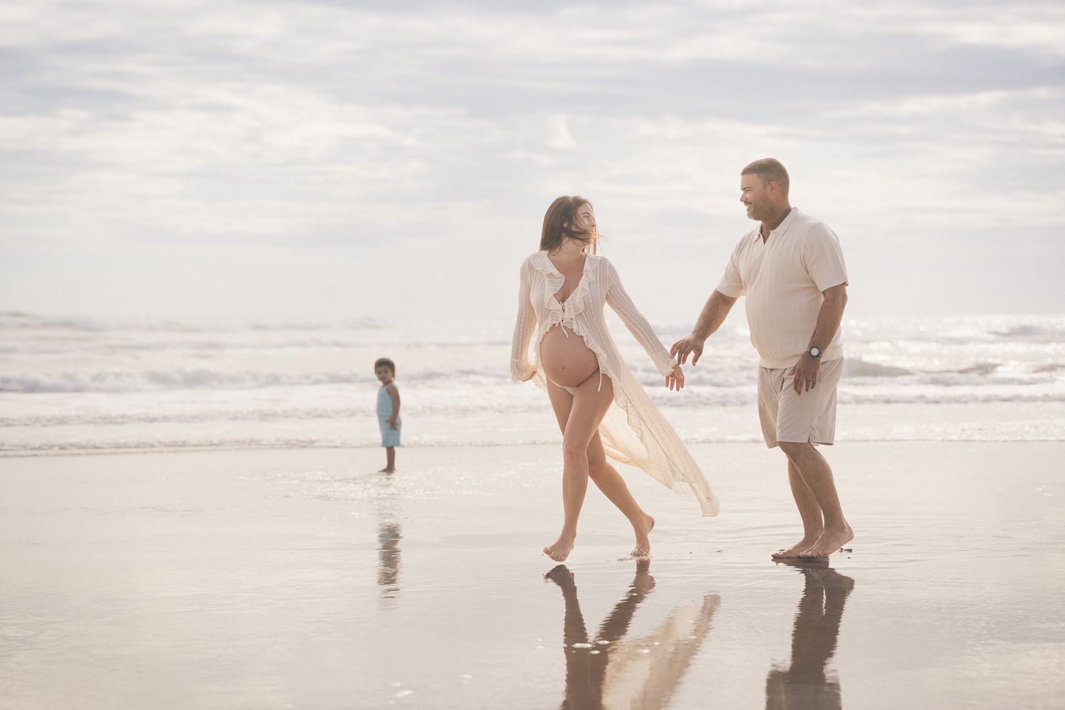 Pregnant couple walking hand in hand on beach with child playing in background.