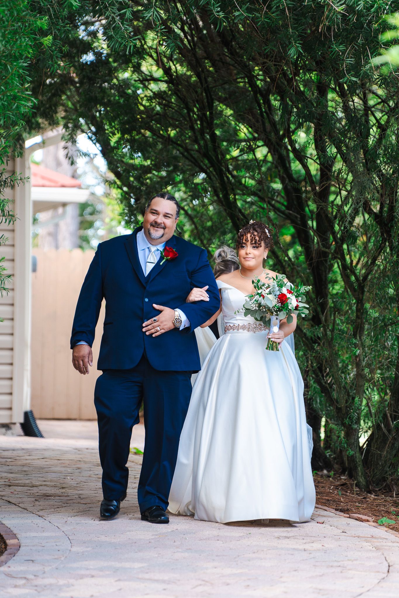 Father of the bride walks daughter in white ball gown through garden pathway to ceremony.