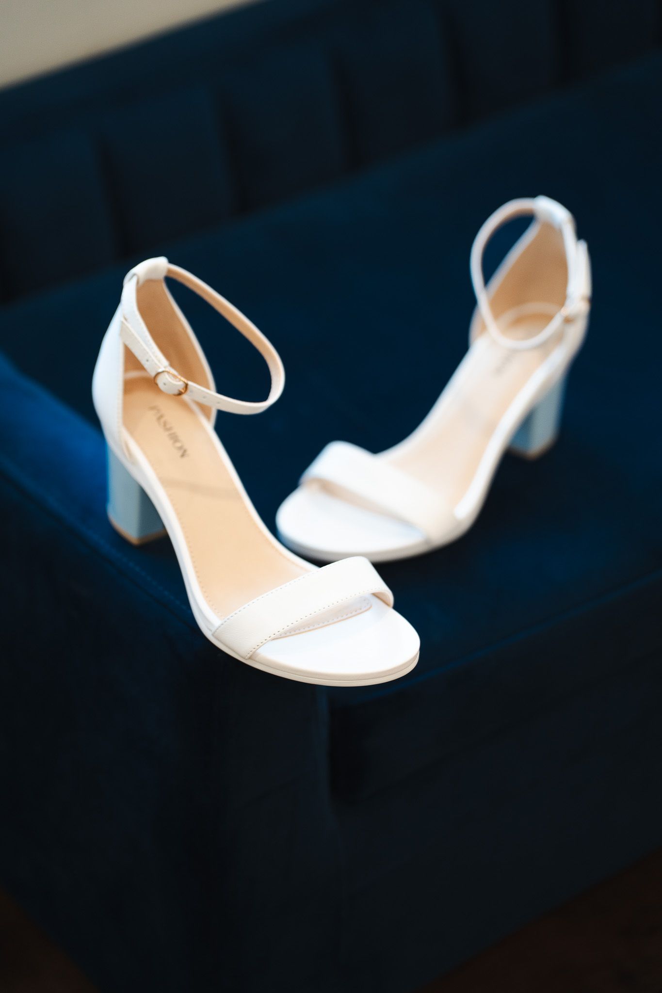 White ankle strap block heel bridal shoes displayed on navy blue velvet chair.