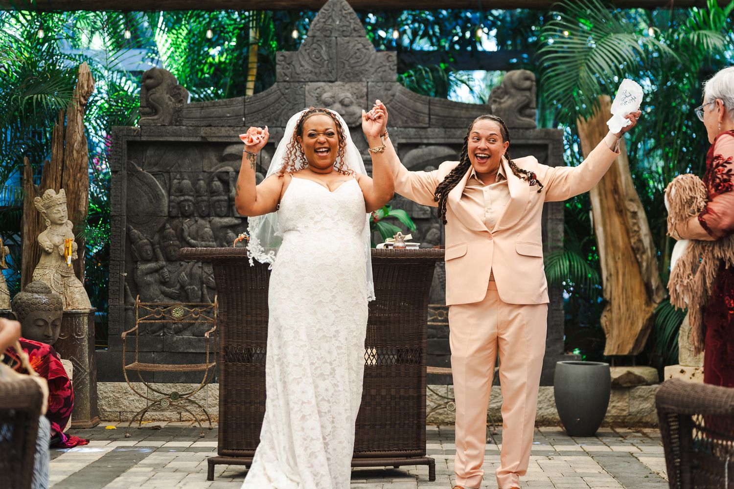 Two brides celebrate joyfully with raised arms after exchanging vows at outdoor wedding.