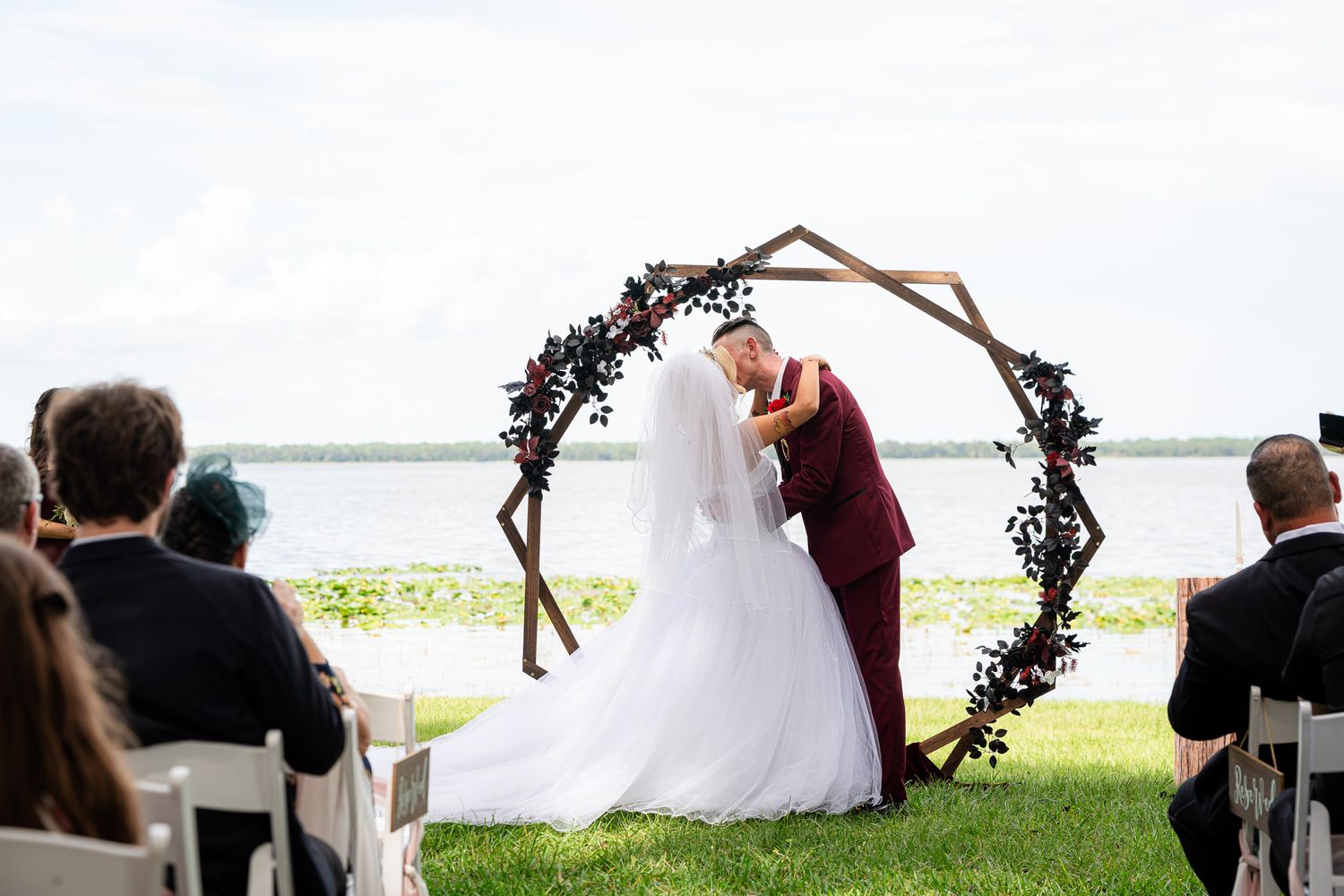 Bride and groom share romantic first kiss under geometric wooden arch decorated with dark floral blooms.