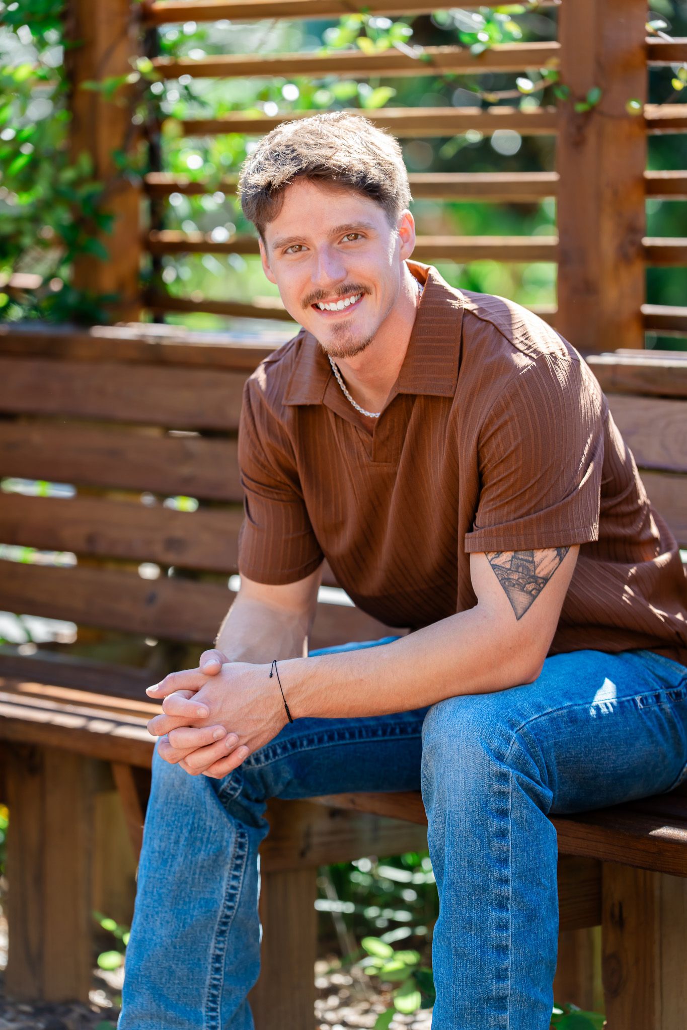 Smiling man with mustache in brown shirt and jeans sitting on wooden bench with green plants behind him.