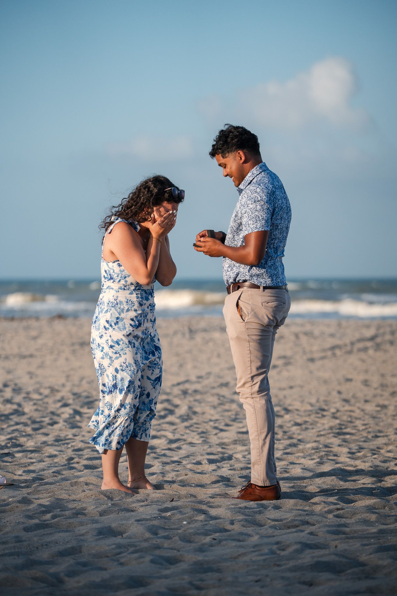 Man presenting a ring box to an emotional woman during a beach marriage proposal at sunset.