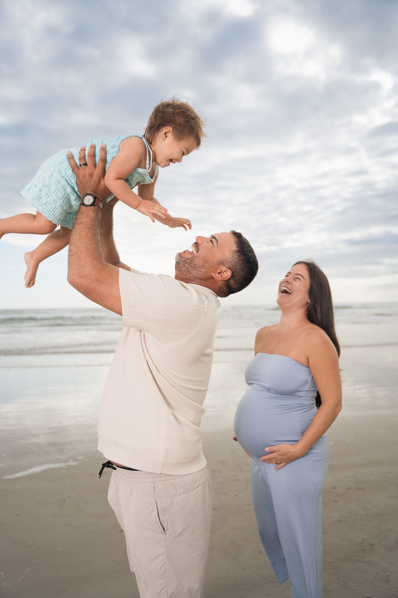 Happy family at the beach as father lifts baby in the air while pregnant mother laughs joyfully beside them.